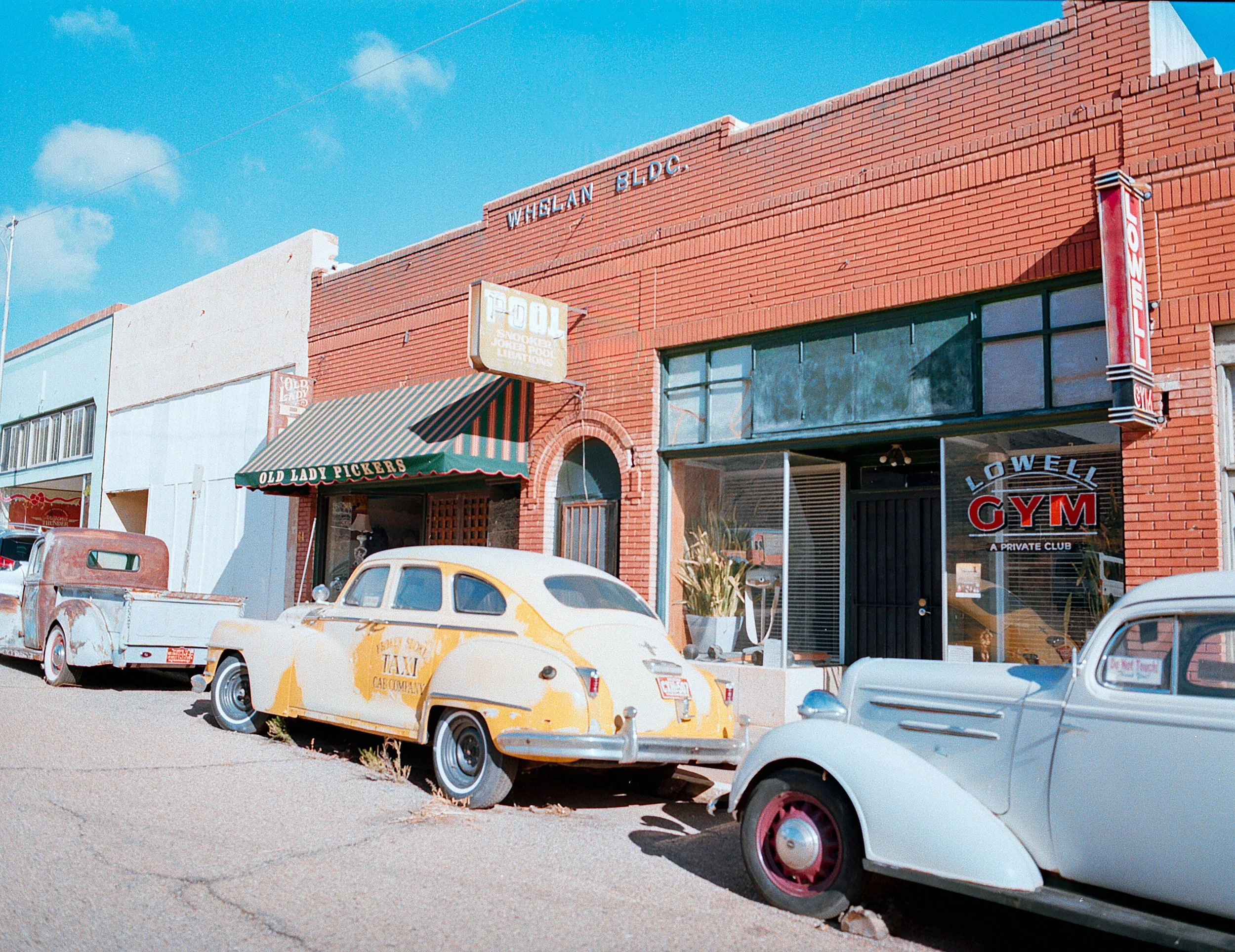 A row of vintage cars parked in front of a brick building with signs for a gym and other businesses, under a blue sky.