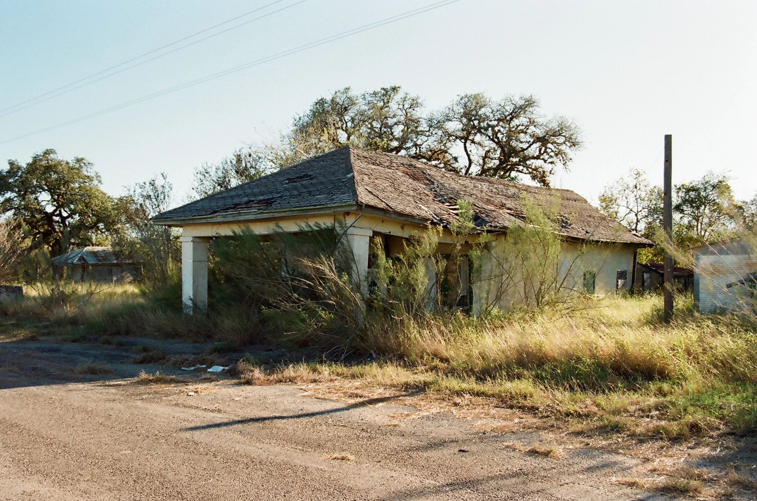 An abandoned house with a damaged roof and overgrown weeds in a rural setting.
