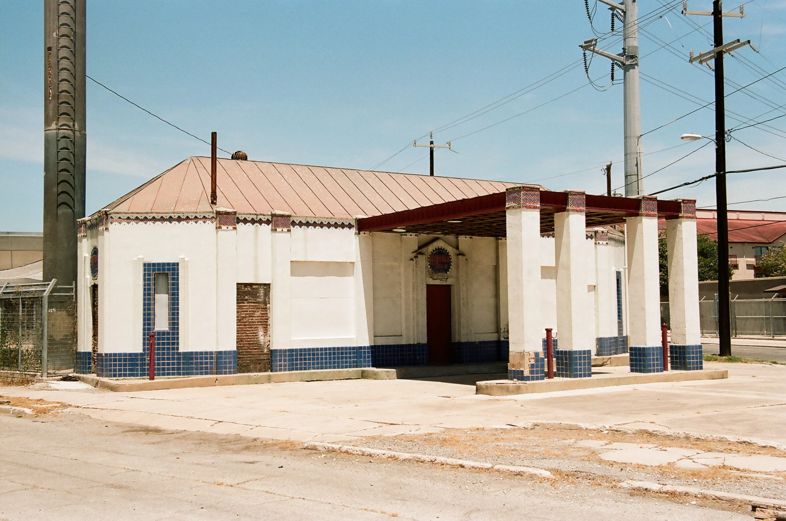 An old, abandoned building with a tiled roof and entrance, featuring white walls with blue tiles at the base, surrounded by utility poles and wires in a mostly empty lot under a clear sky.