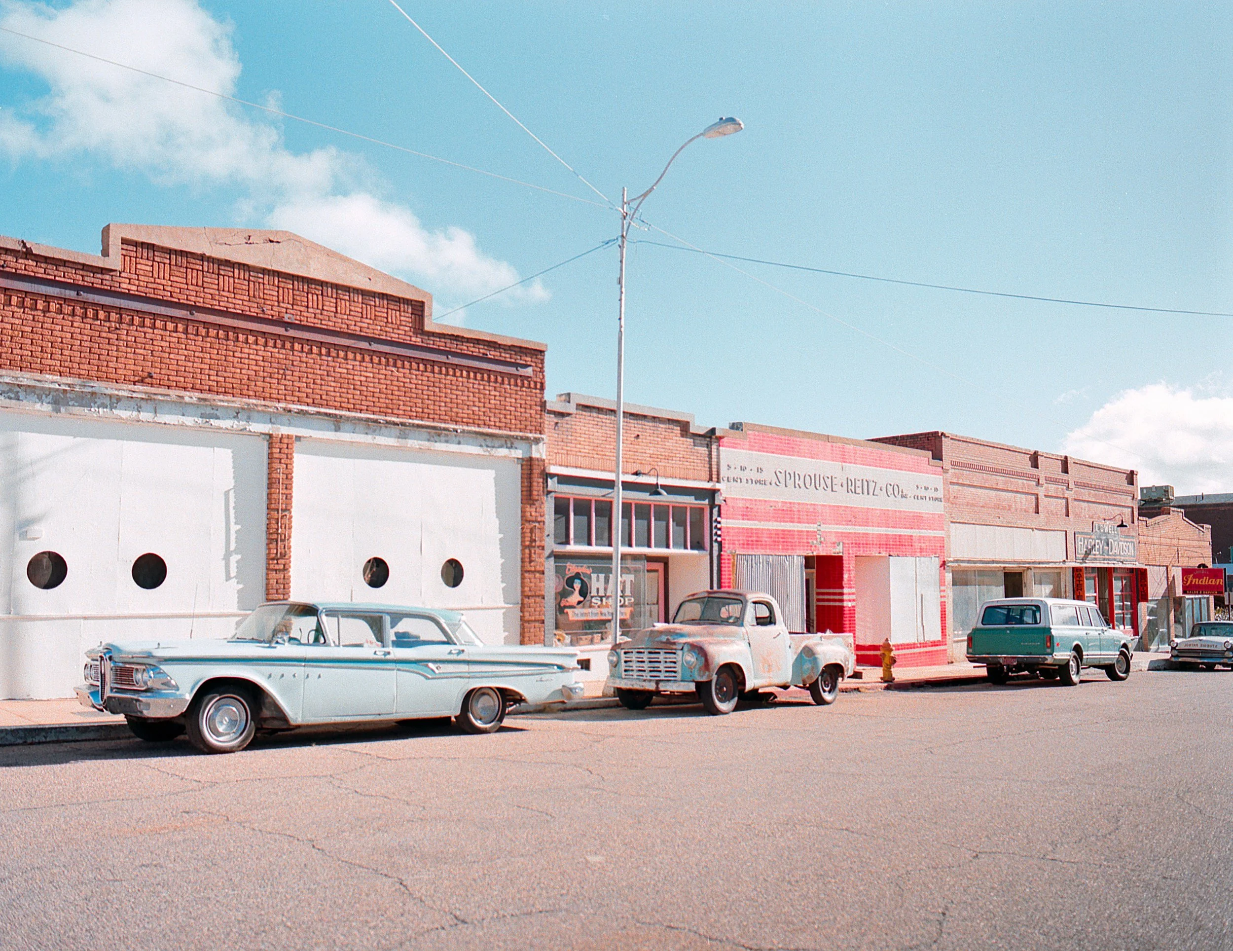 Street scene with vintage cars parked in front of brick and painted storefronts on a sunny day.