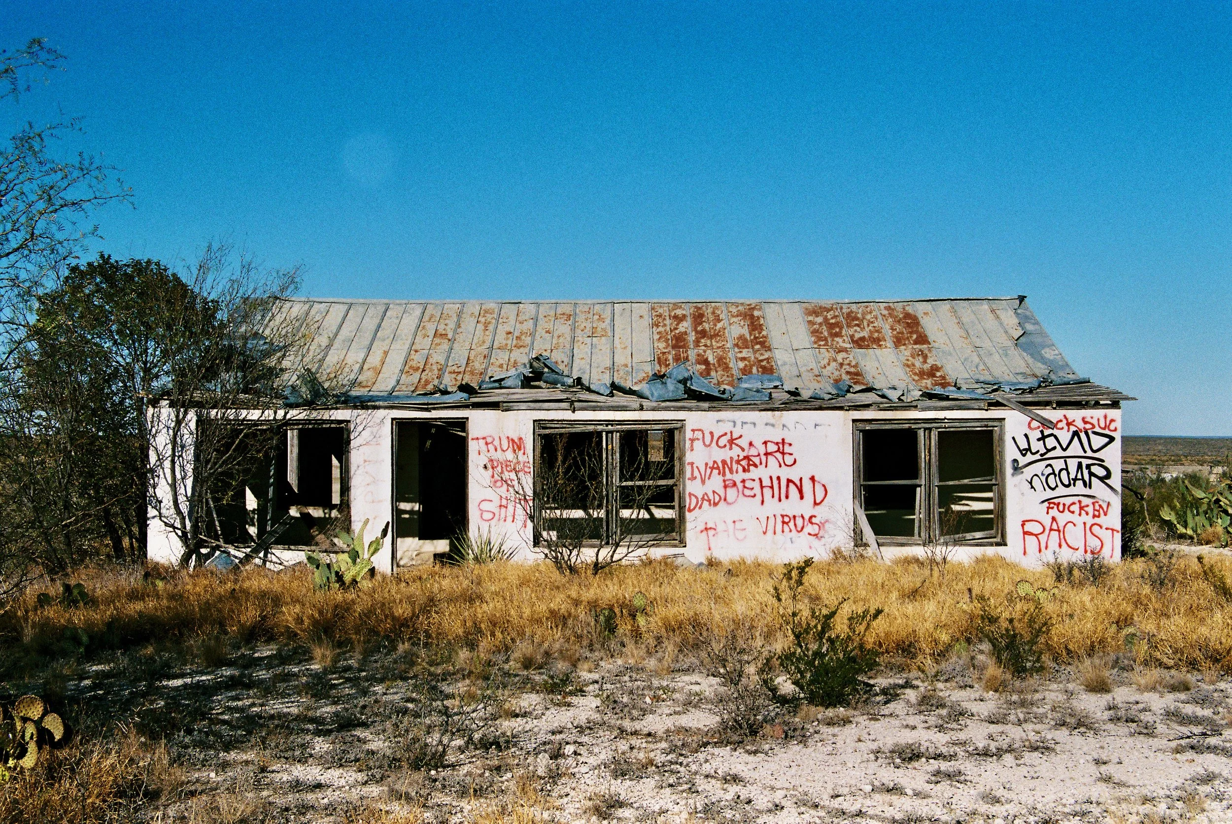Abandoned house in a desert landscape with graffiti on the walls, including messages like 'F*** THE VIRUS', 'F*** RACIST', and other obscenities, with a rusted metal roof and overgrown dry grass in the foreground.