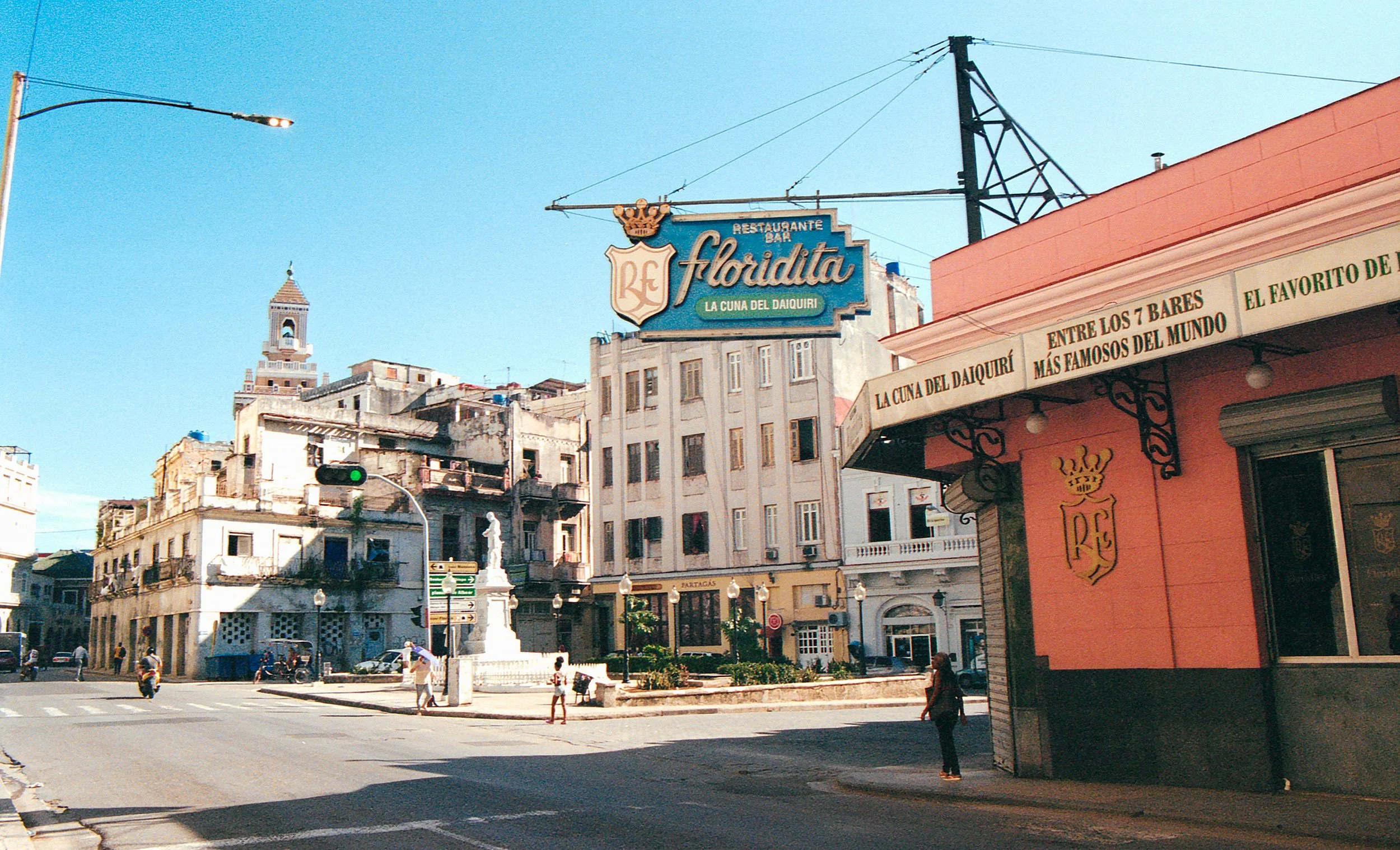 City street scene in an urban area with old buildings, a church tower in the background, a traffic light, and a corner restaurant sign for 'Floridita' with a terrace and people walking.
