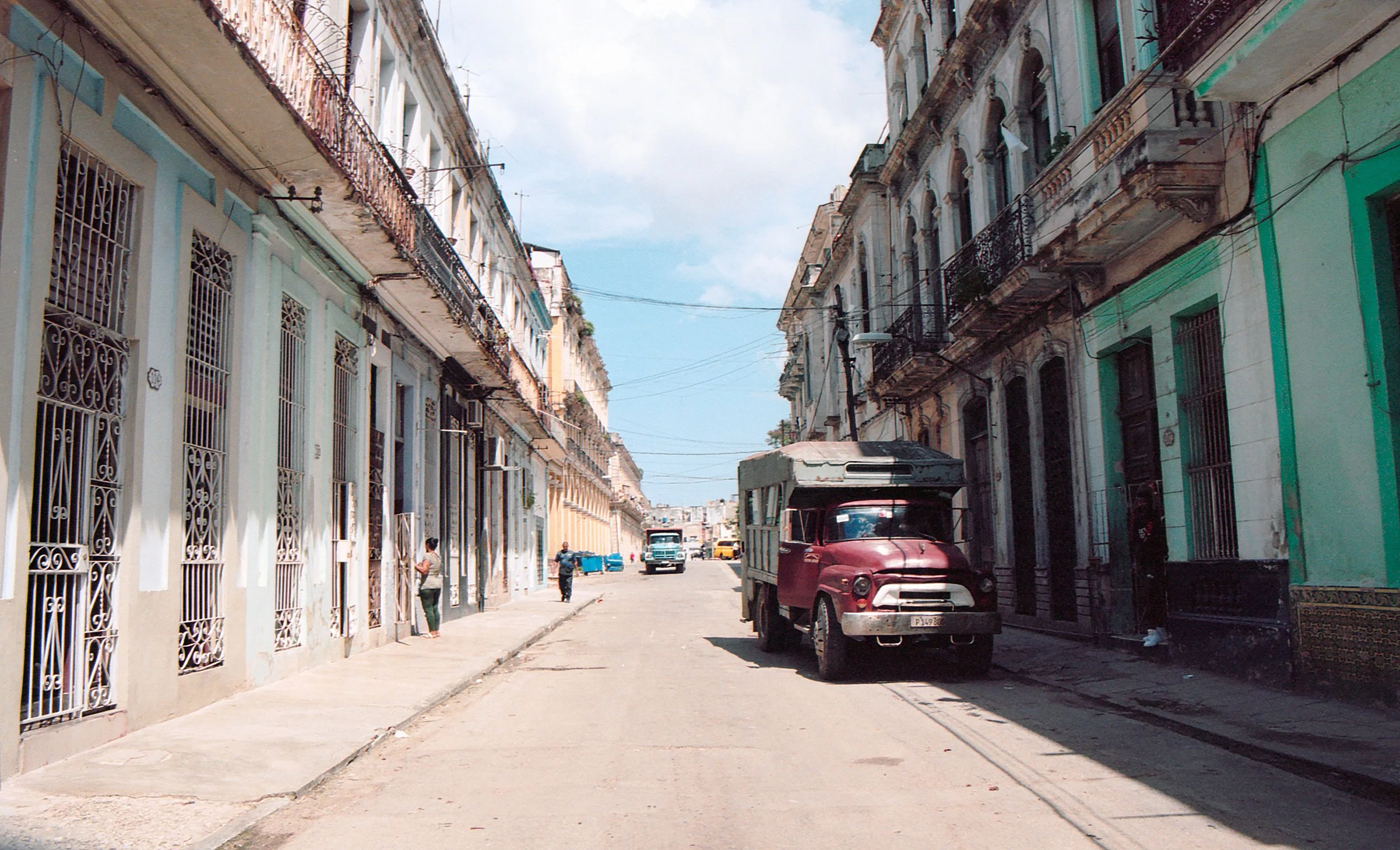 A street scene with colorful old buildings, an old red truck parked on the side, and a few people walking along the sidewalk.