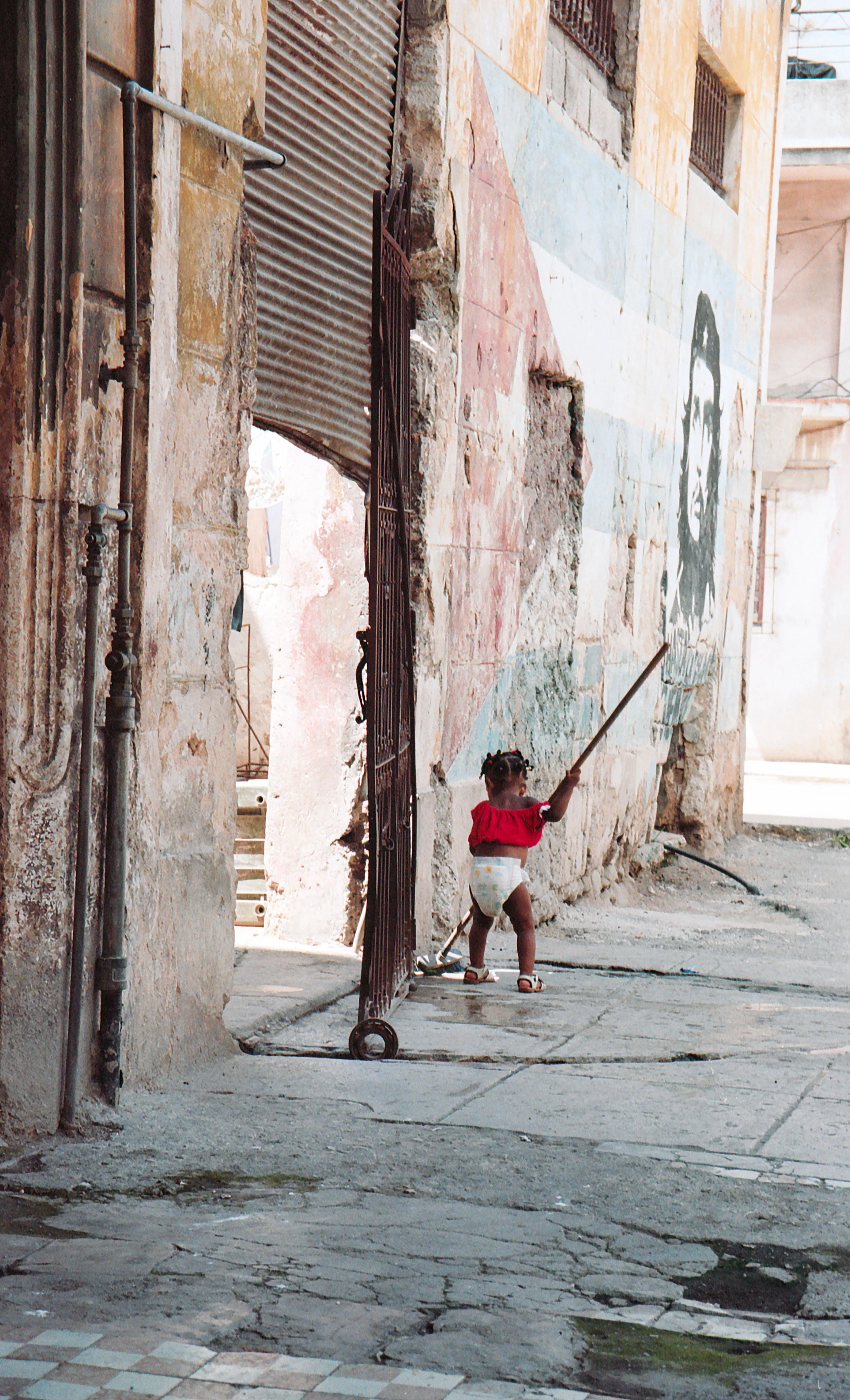 A young girl in a red top and diaper plays with a stick near an open gate on a street with weathered buildings, one of which has a mural of a face on the wall.