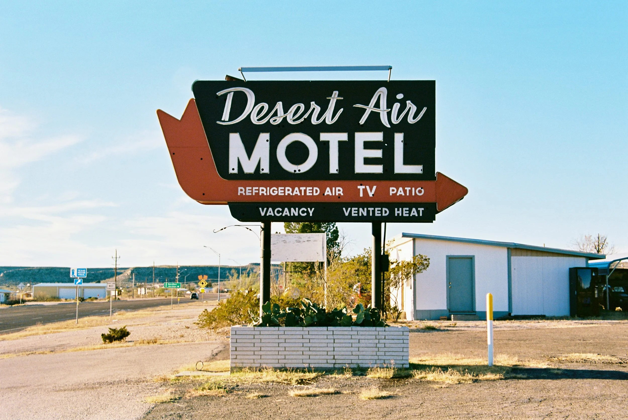 Sign for Desert Air Motel with amenities listed as refrigerated air, TV, patio, vacancy, and vented heat, situated in a desert area with a small building nearby.