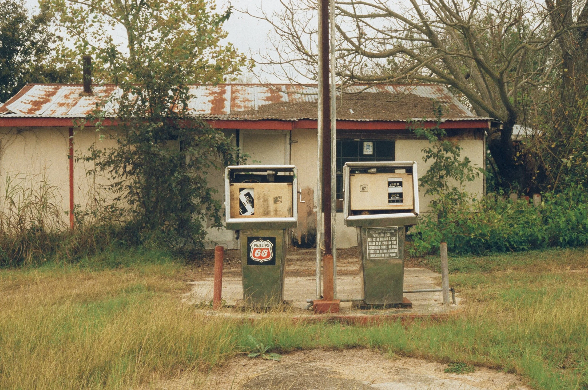 Two old, rusty gasoline pumps in front of a weathered house with a rusted metal roof, surrounded by overgrown grass and trees.