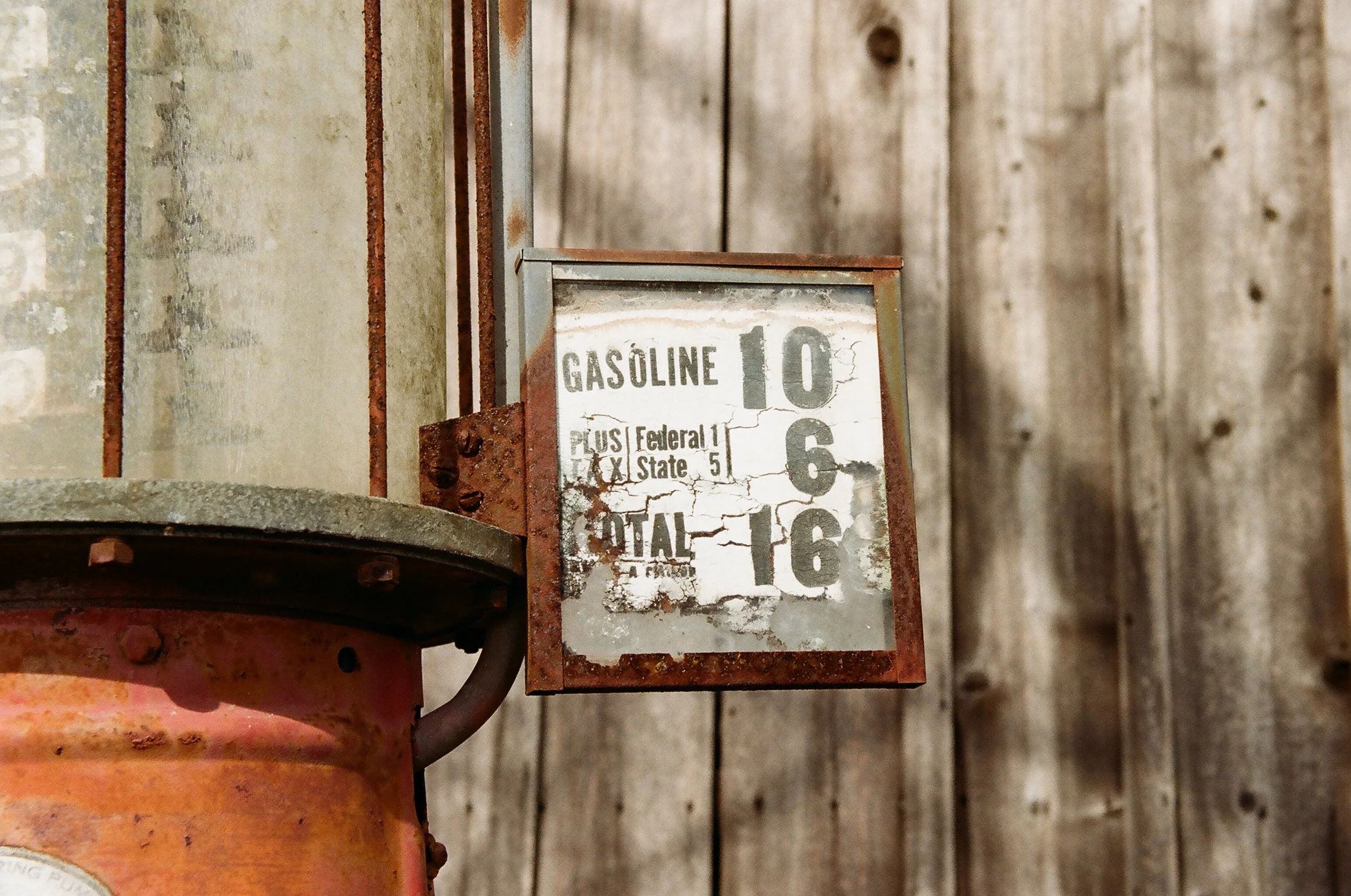 Rusty old gasoline pump with a weathered price sign displaying fuel costs, against a wooden wall background.