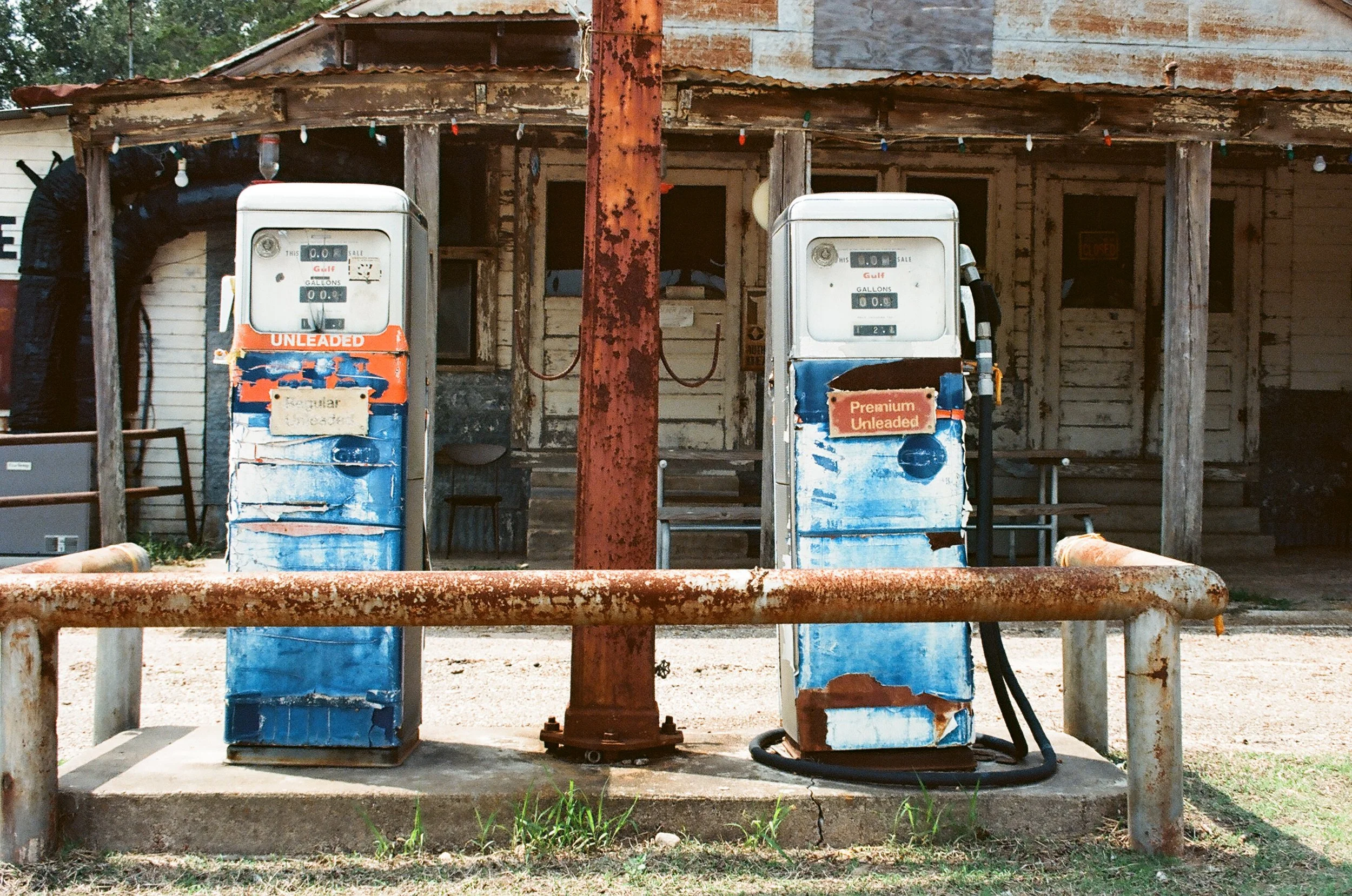 Two old rusty gas pumps in front of a weathered wooden building, with a rusty safety barrier and string lights overhead.