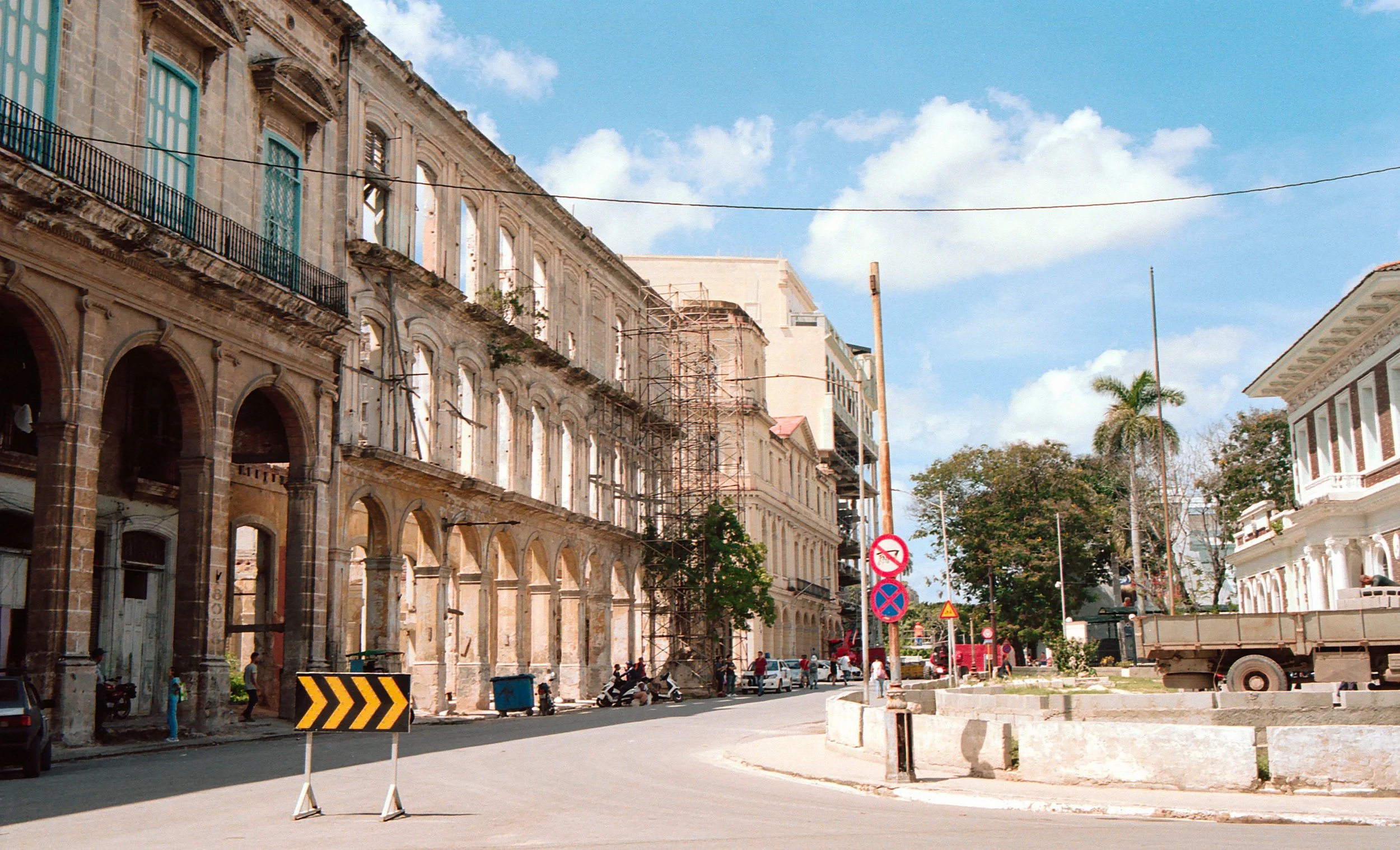 Street scene with old buildings under renovation, scaffolding, parked motorcycles, cars, and a truck, with signs indicating no parking and no entry, in a sunny weather with blue sky and scattered clouds.