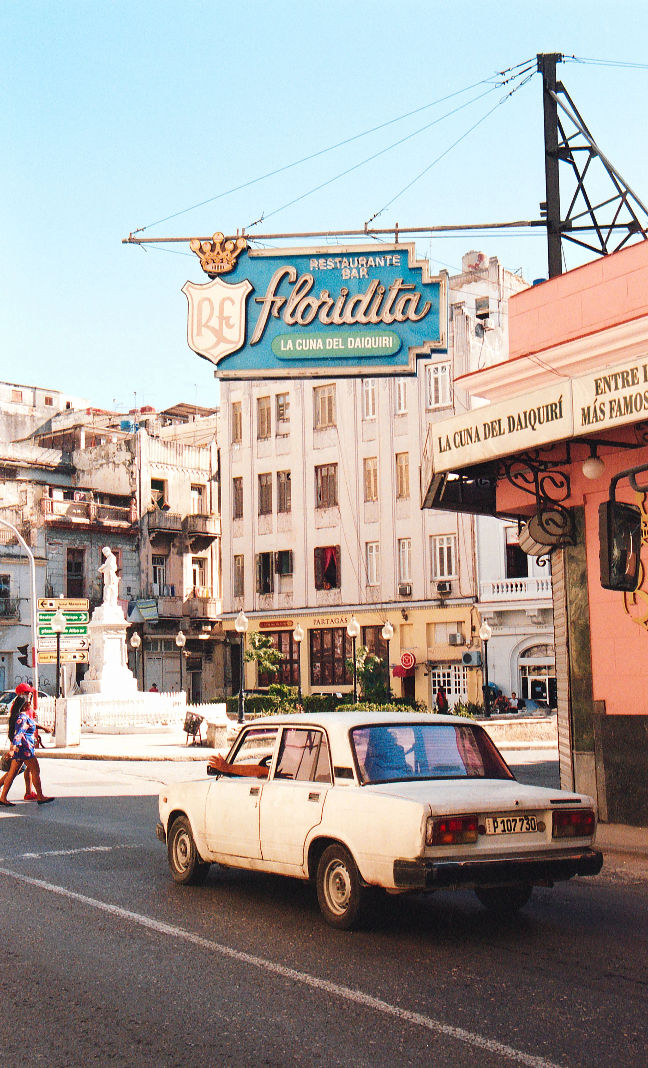 A street scene with a white vintage car and a pink building with an sign that reads 'Florida' and 'La Cuna del Daiquiri', with buildings and a statue in the background.