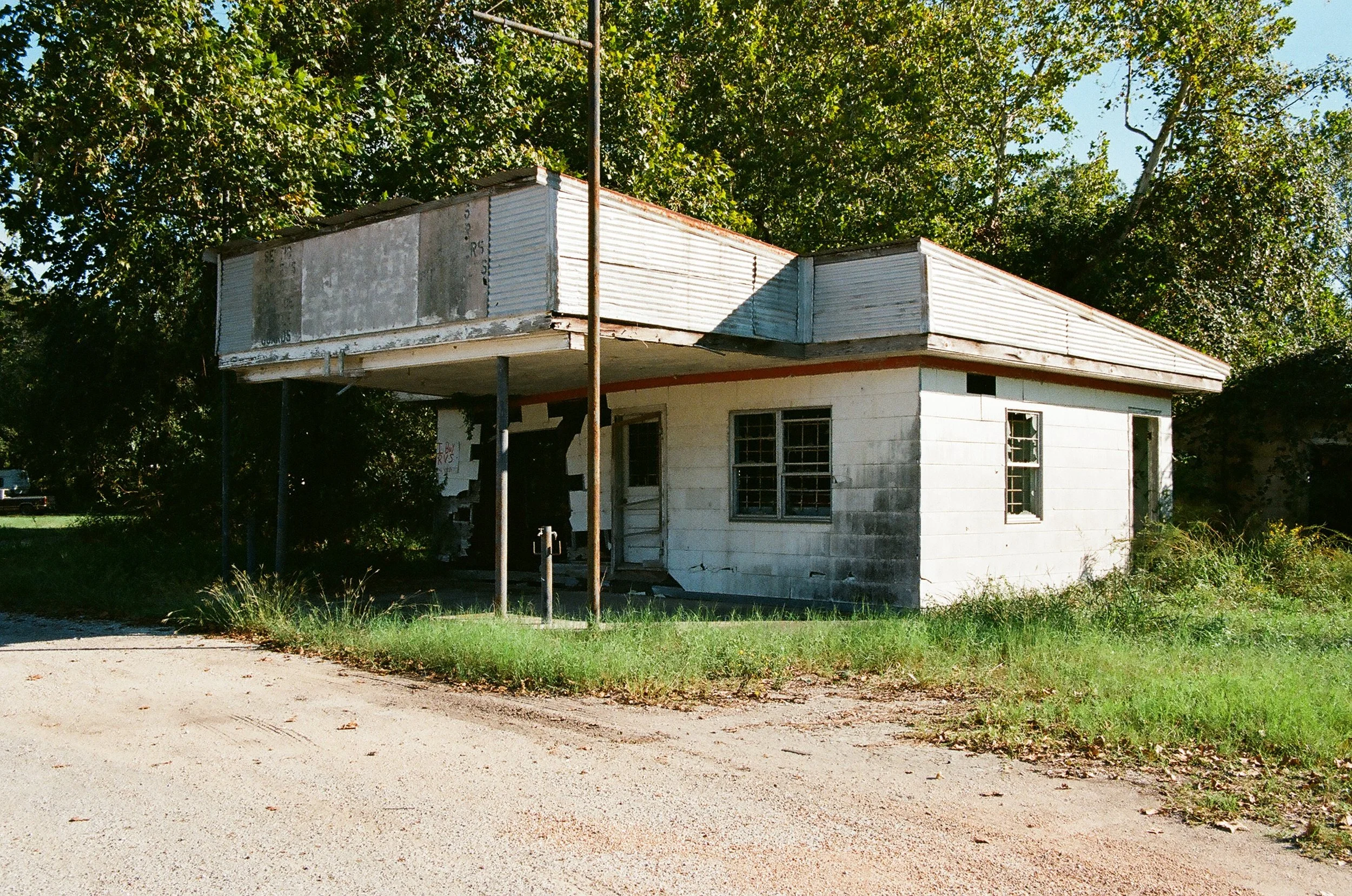 Old, abandoned house with a damaged exterior, overgrown grass, and a rusty pole in front.