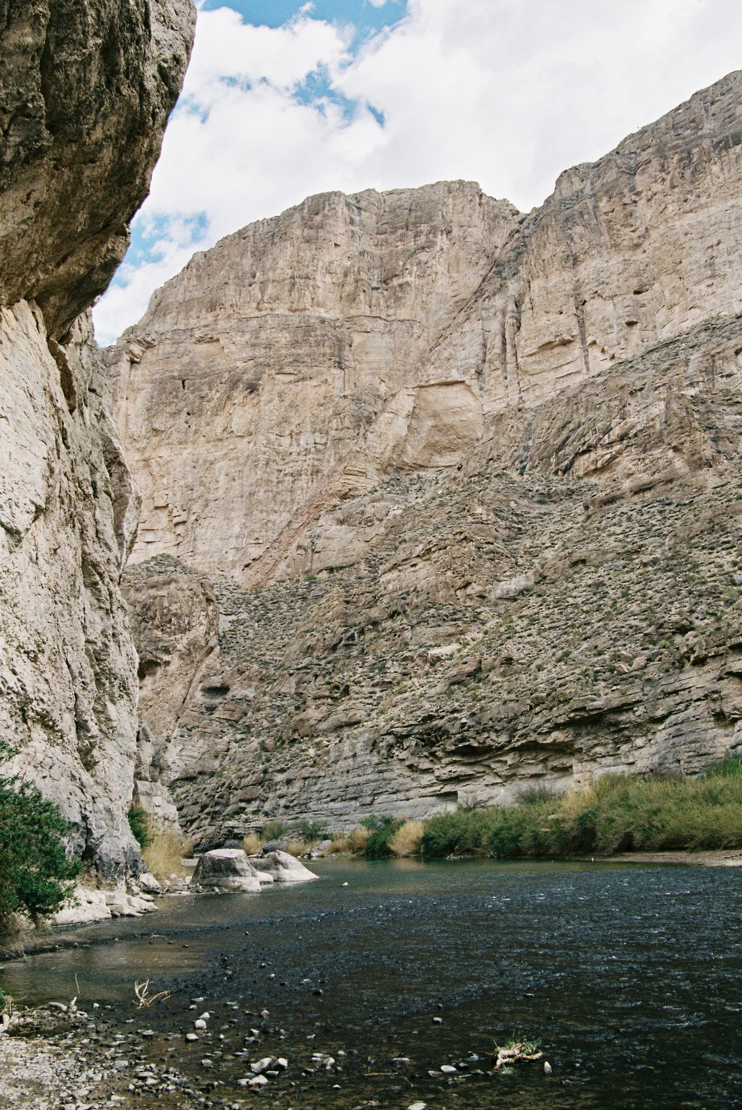 A narrow river runs through a deep canyon with tall, rocky, beige-colored cliffs on both sides and some sparse green vegetation along the water's edge, under a partly cloudy sky.