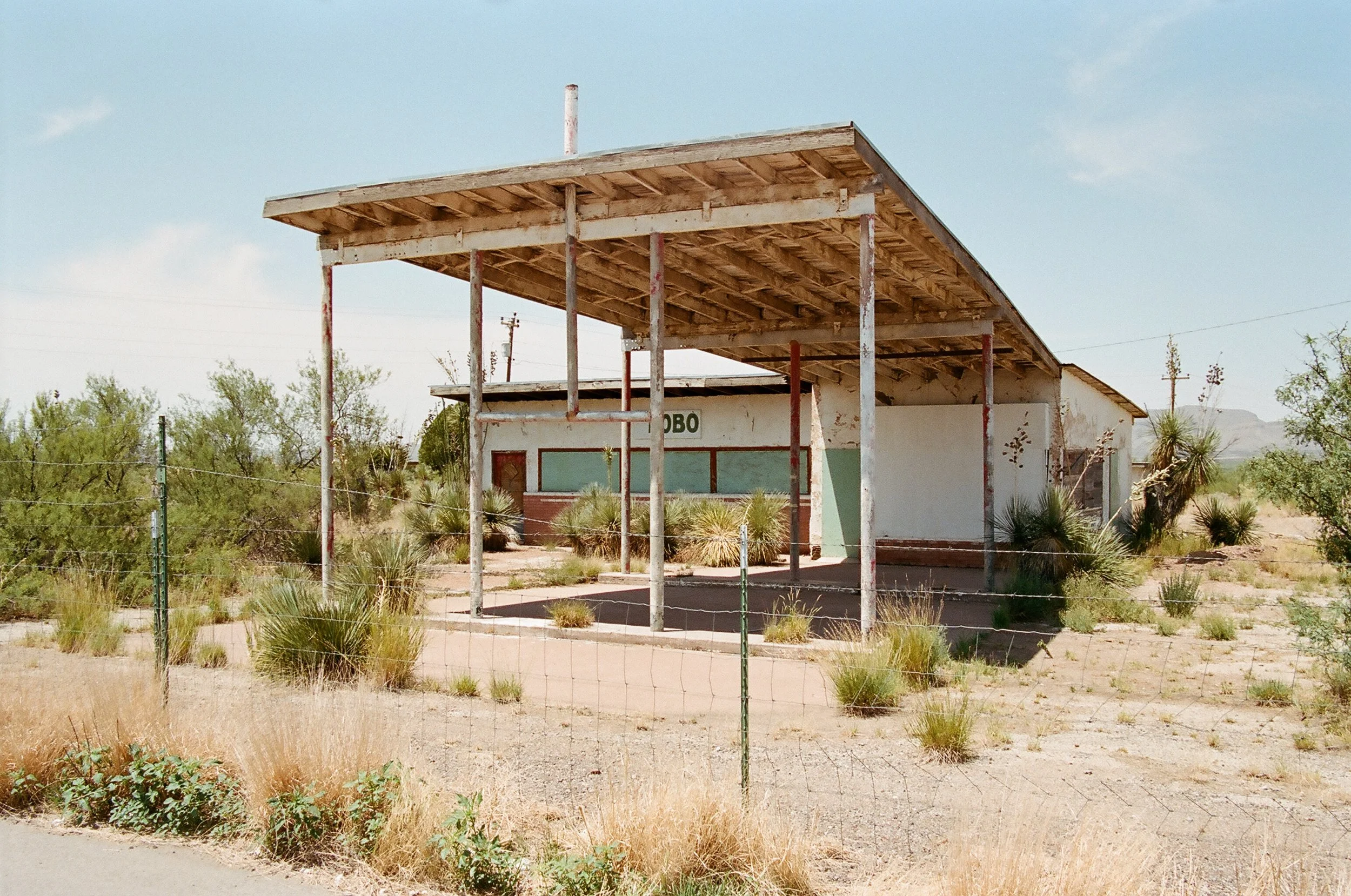 An abandoned building in a desert landscape with a large wooden awning, surrounded by dry bushes and sparse desert vegetation.