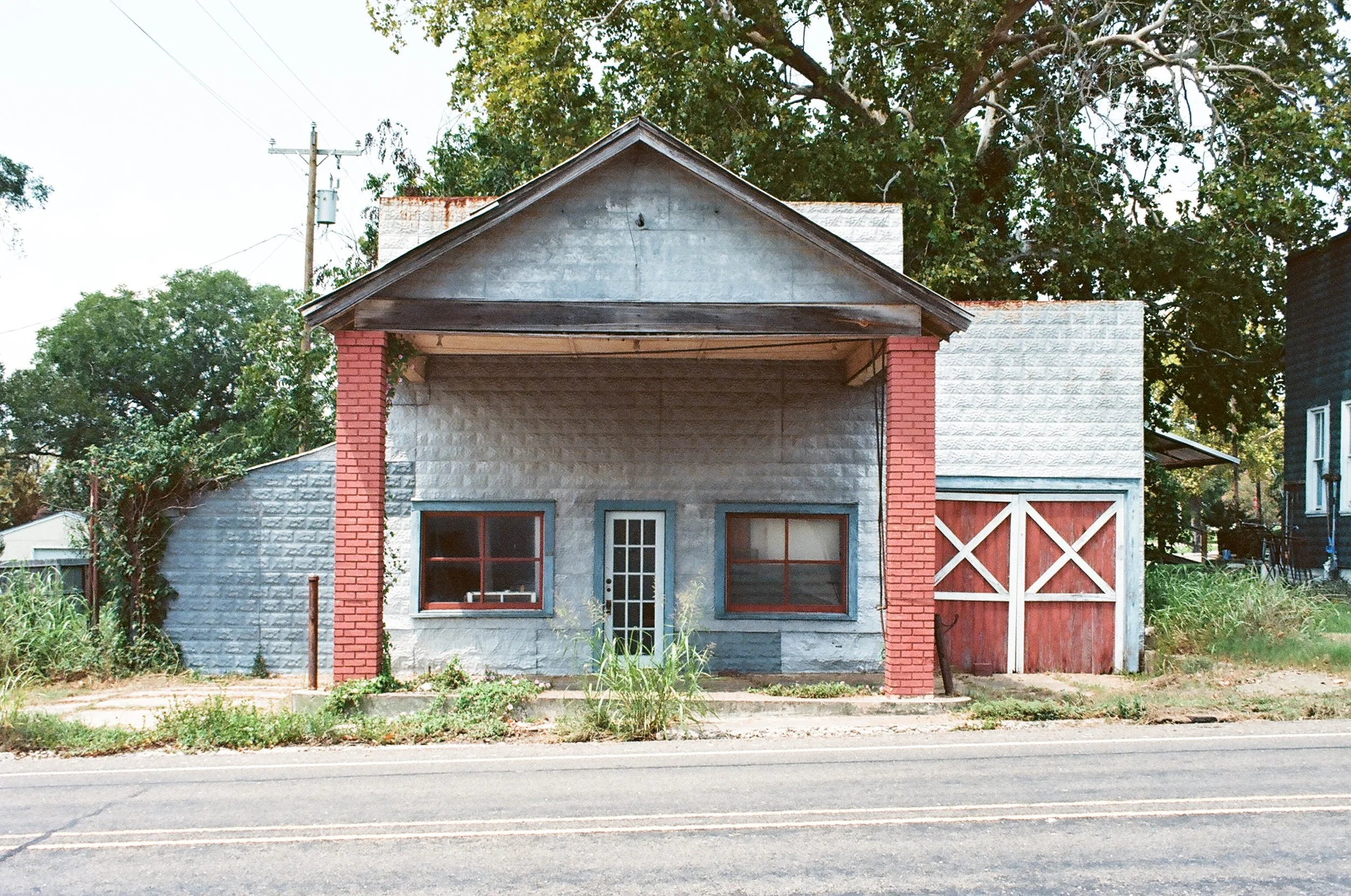 Small house with gray walls, red-brick columns, and a wooden roof, located on a street with trees in the background.