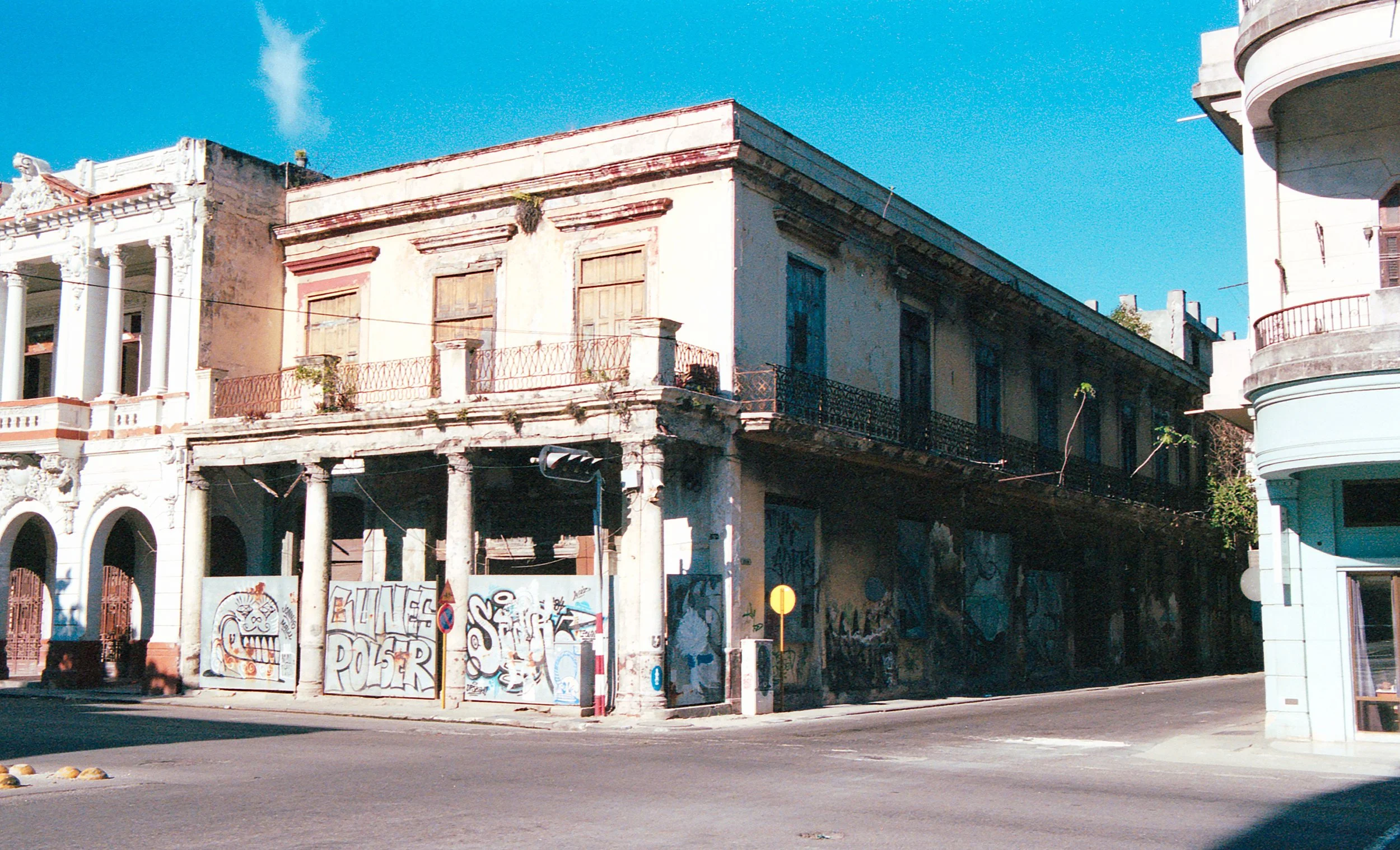 Old, weathered building with graffiti and graffiti-covered walls, next to a white building with rounded balconies, under a clear blue sky.