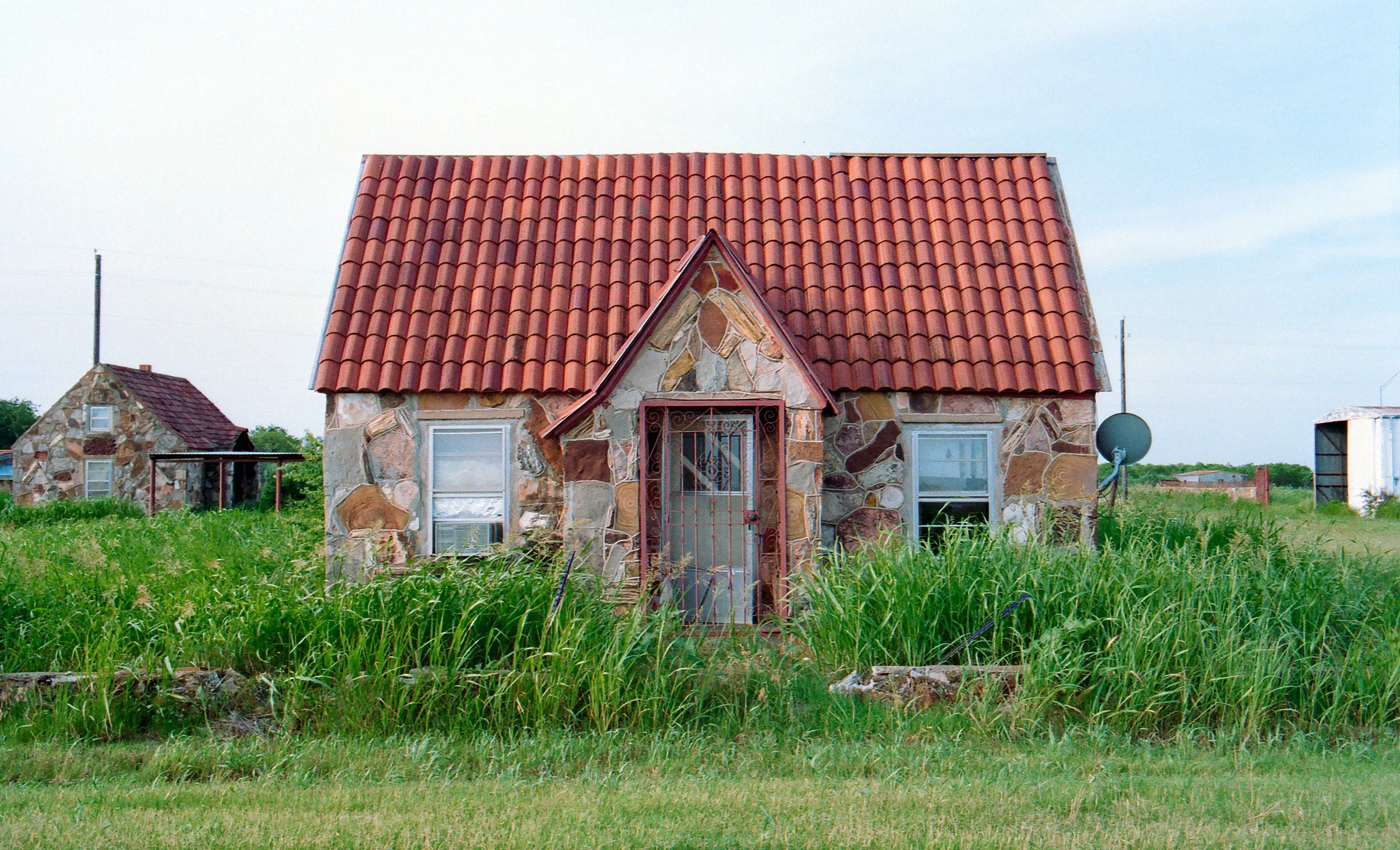 A small stone cottage with a red tiled roof, surrounded by tall grass and a lush green lawn.