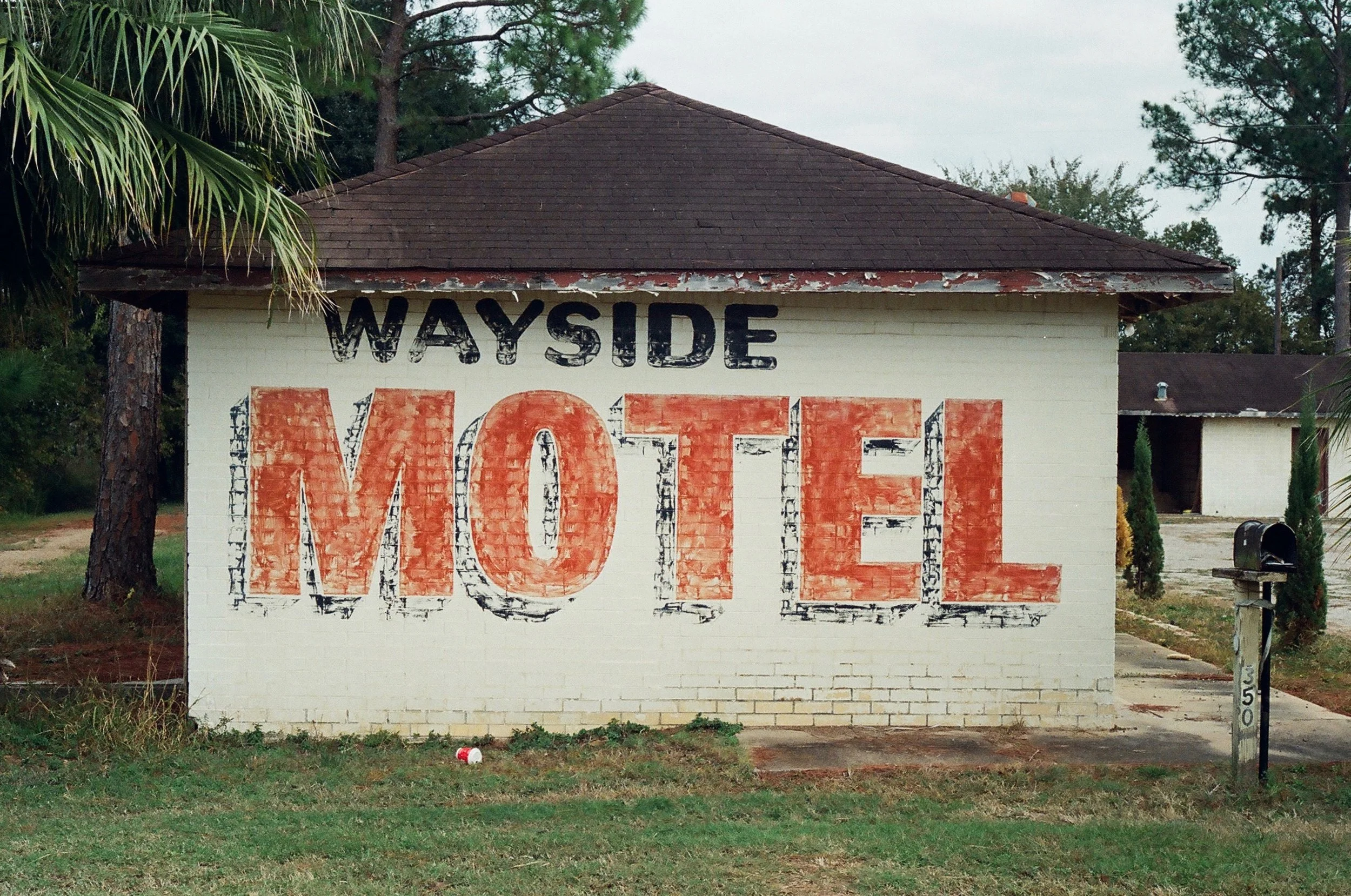 A white brick house with a mural that says 'WAYSIDE MOTEL' painted on the front wall. The word 'WAYSIDE' is in black, and 'MOTEL' is in large, worn-looking orange and black letters. There are trees and neighboring buildings in the background.