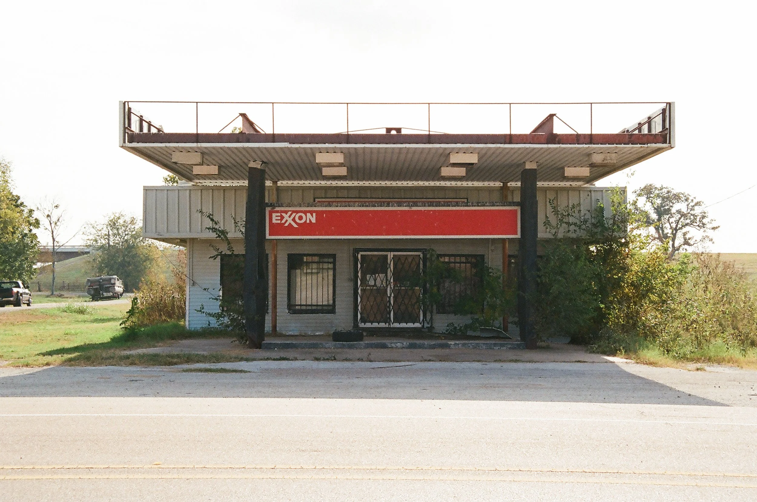 Empty roadside gas station with an Exxon sign, plants growing around the building, and a balcony on the upper level.