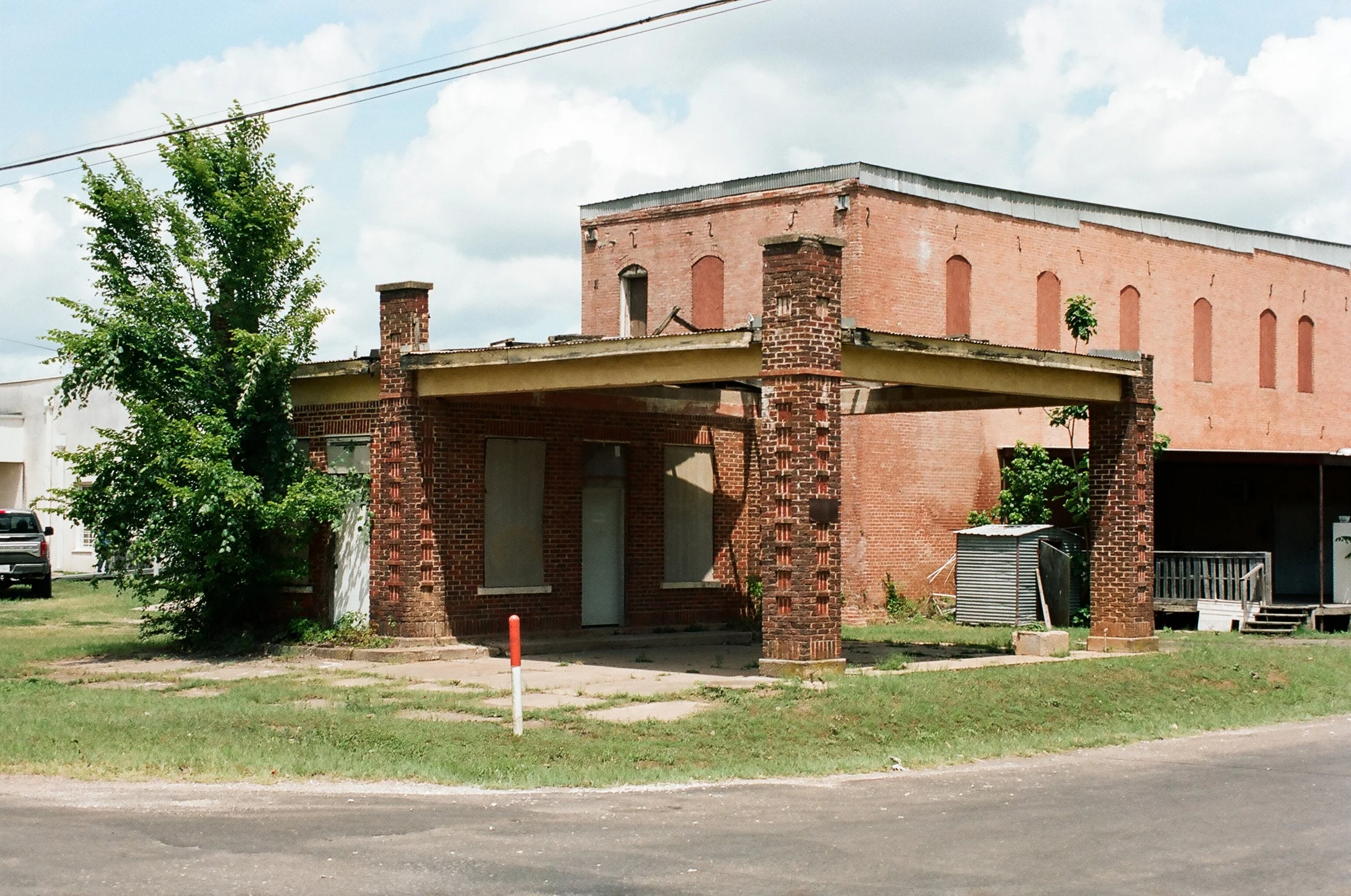 An old brick building with boarded-up windows, a small porch, and a tree on the left side under a cloudy sky.