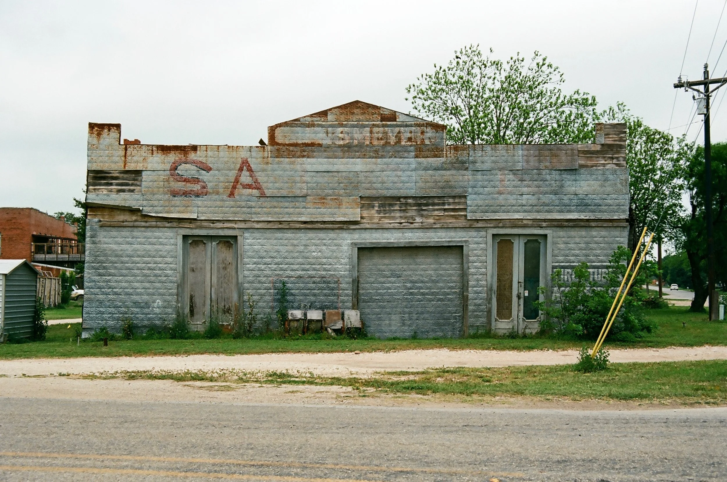 An old, abandoned building with a weathered metal facade, partially covered with rust and dirt, with faded letters 'SA' on the upper left, and boarded-up doors and windows. There are weeds and two yellow poles leaning against a bush to the right.