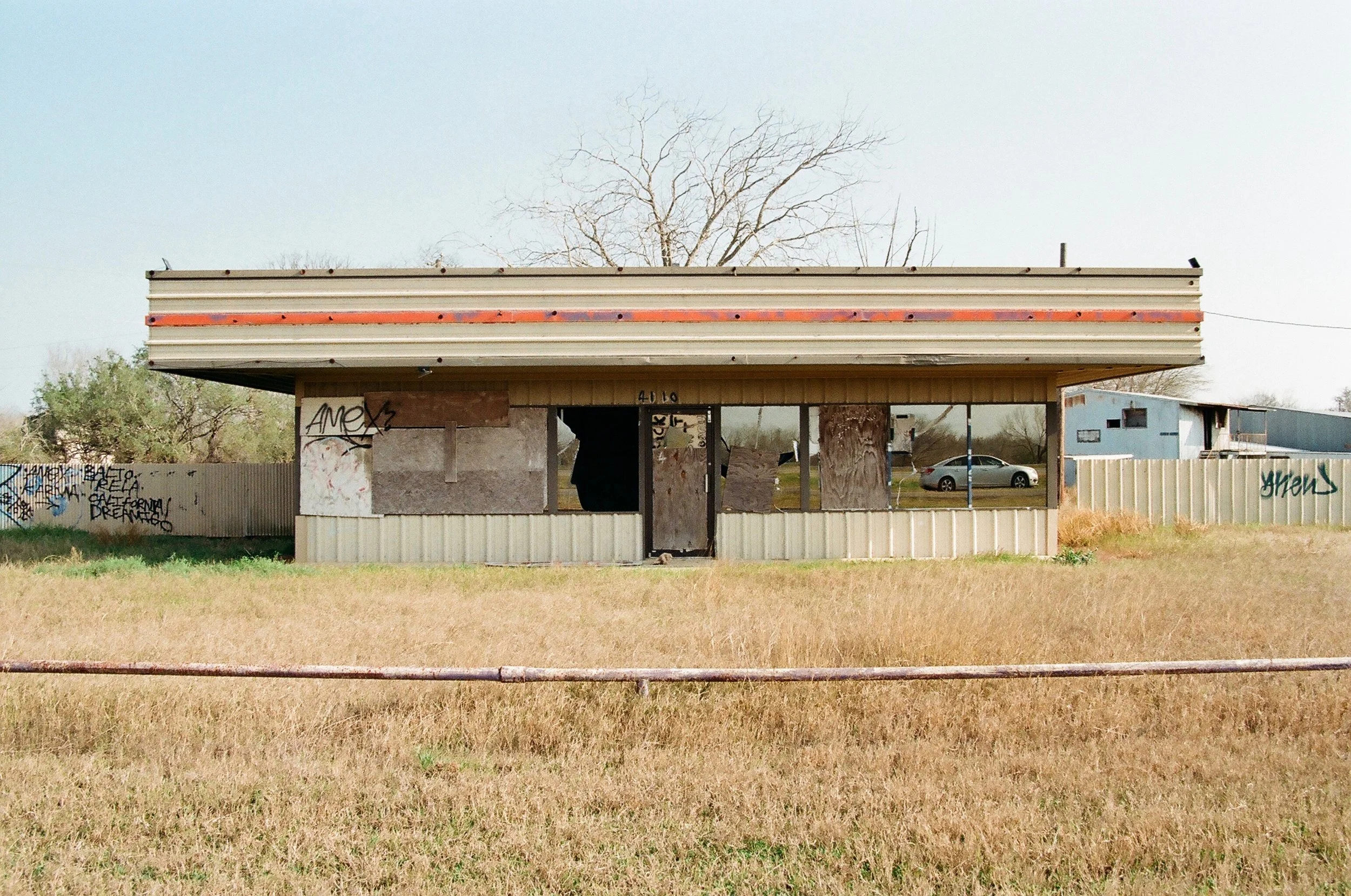 An abandoned, boarded-up building with graffiti, situated in a grassy field with a car and some blue structures in the background.