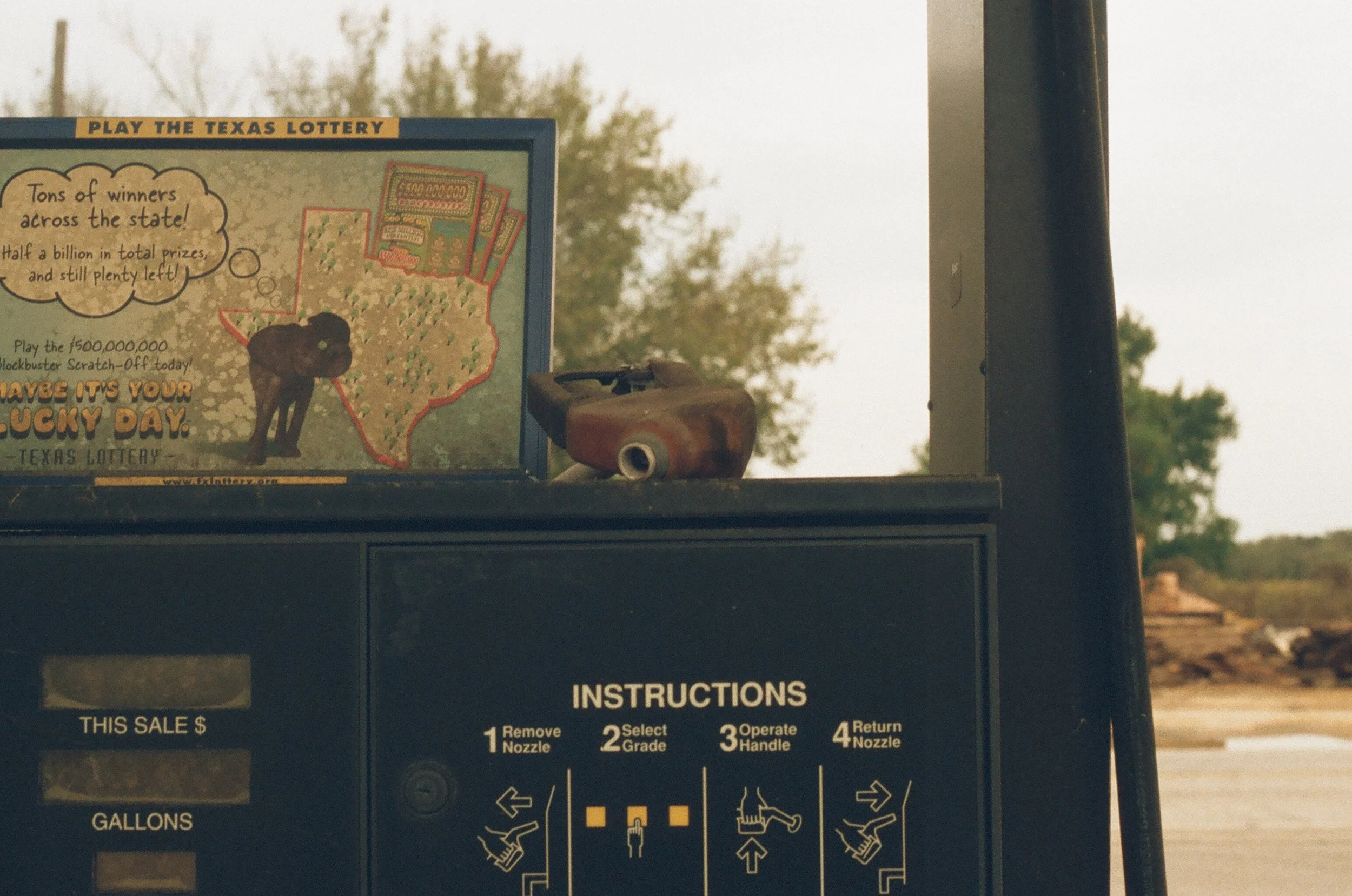 A lottery ticket vending machine with a sign encouraging playing the Texas Lottery and a rusty handheld gas pump with instructions on how to operate it.