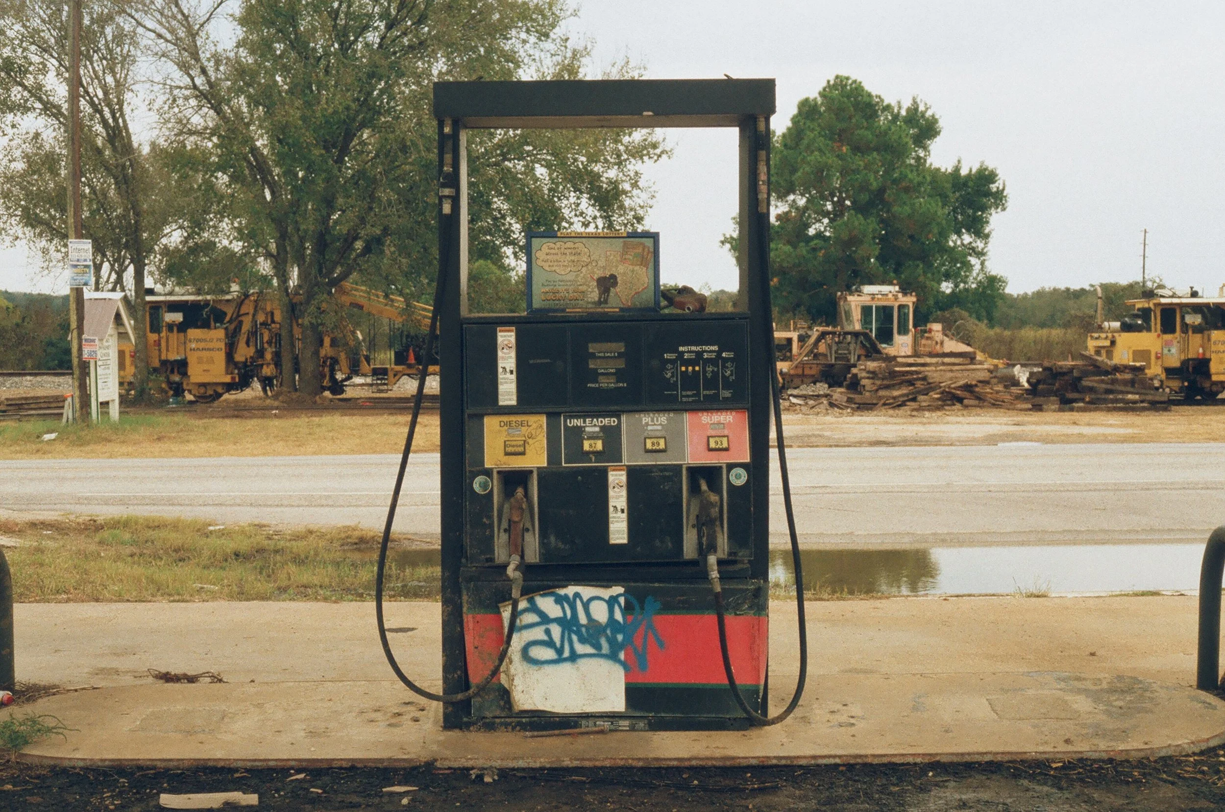 A gas pump at a deserted fuel station with graffiti on the lower part, and construction equipment and trees in the background.