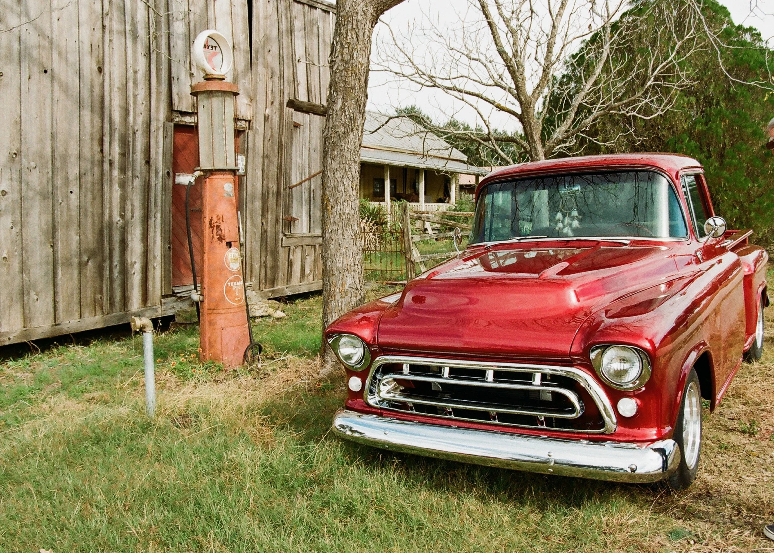 A vintage red pickup truck parked on grass beside an old wooden building with a weathered gas pump nearby.