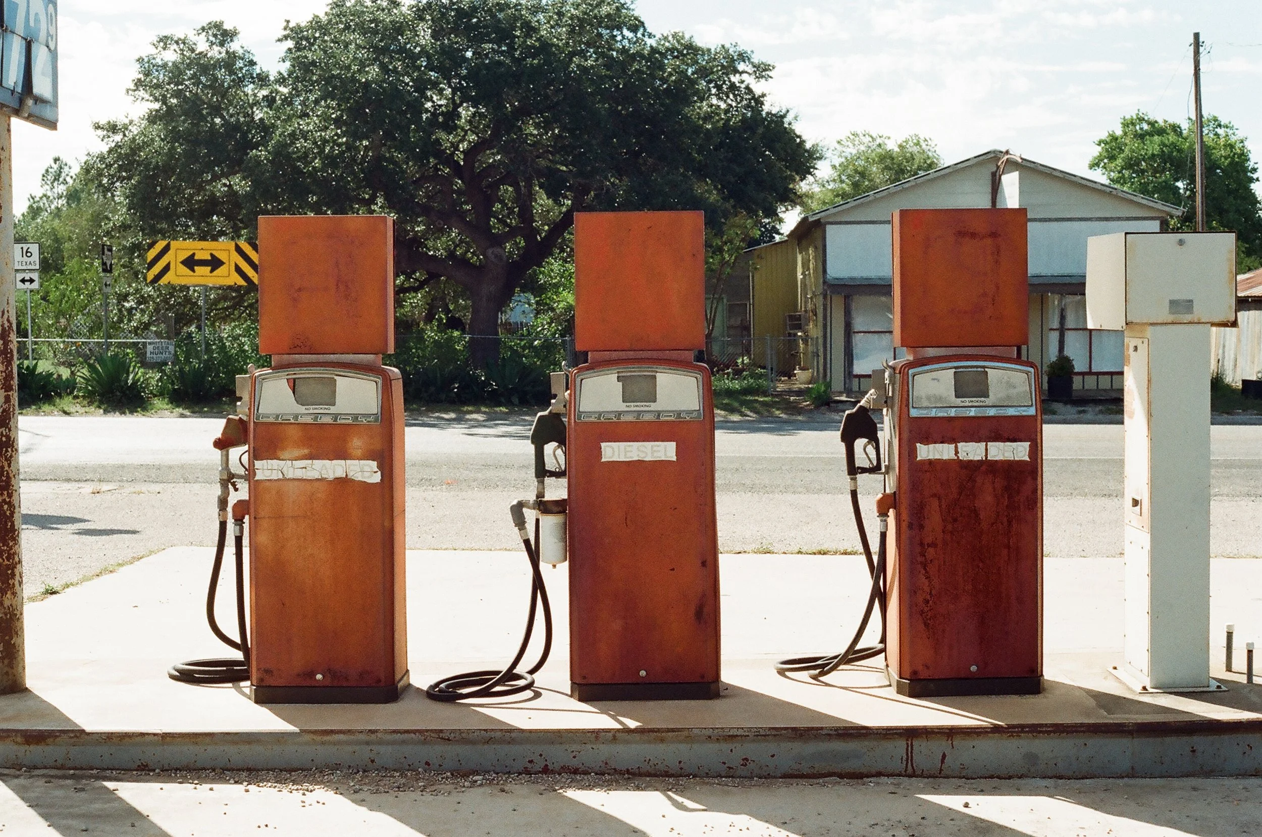 Old, rusty gas pumps at a deserted gas station with a small house and trees in the background.