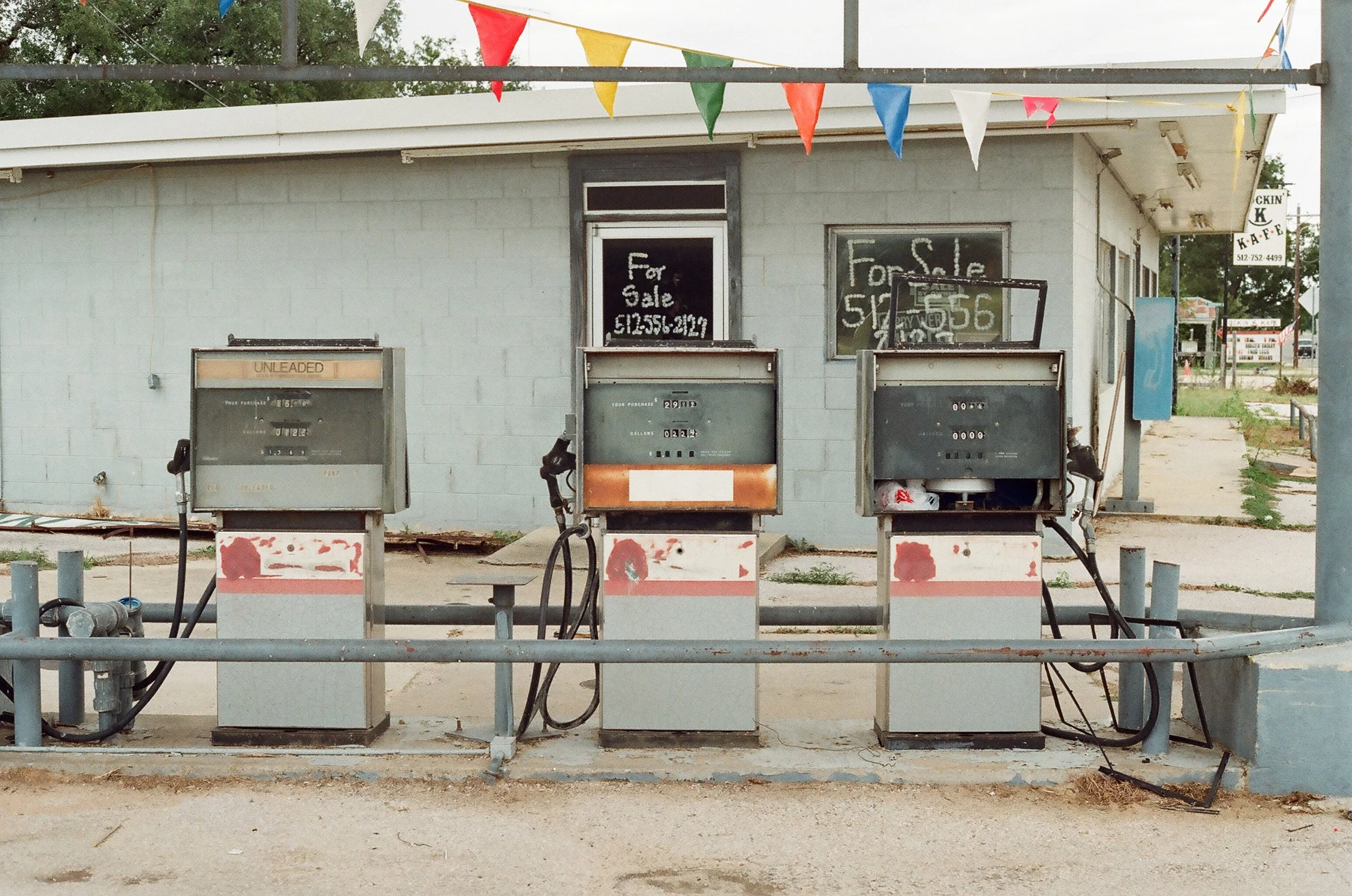 Three old fuel pumps in front of a light blue building with for sale signs on the windows, and colorful triangular flags hanging overhead.