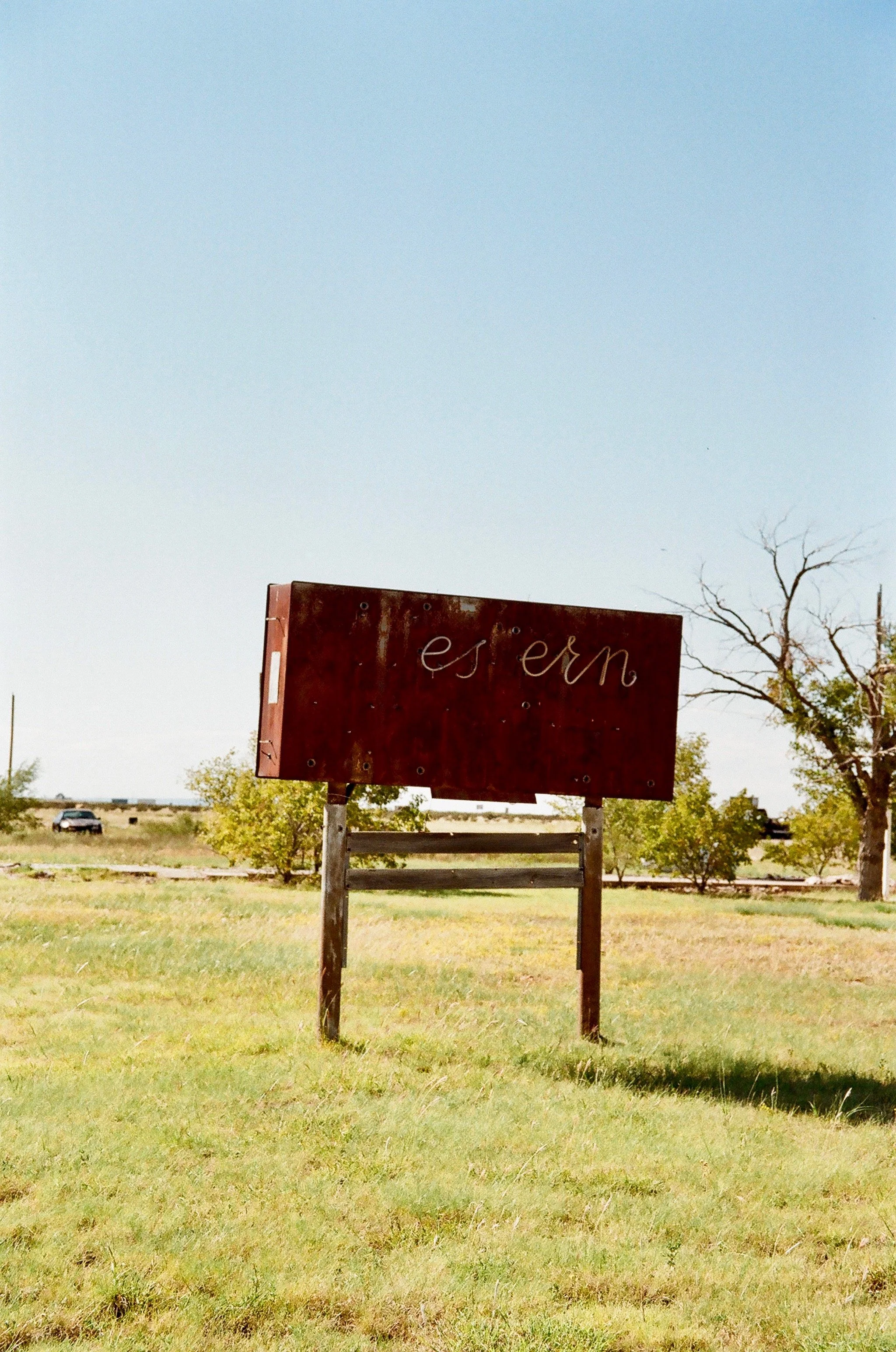 Rusted sign with the word 'es em' in cursive on a grassy field under a clear sky.