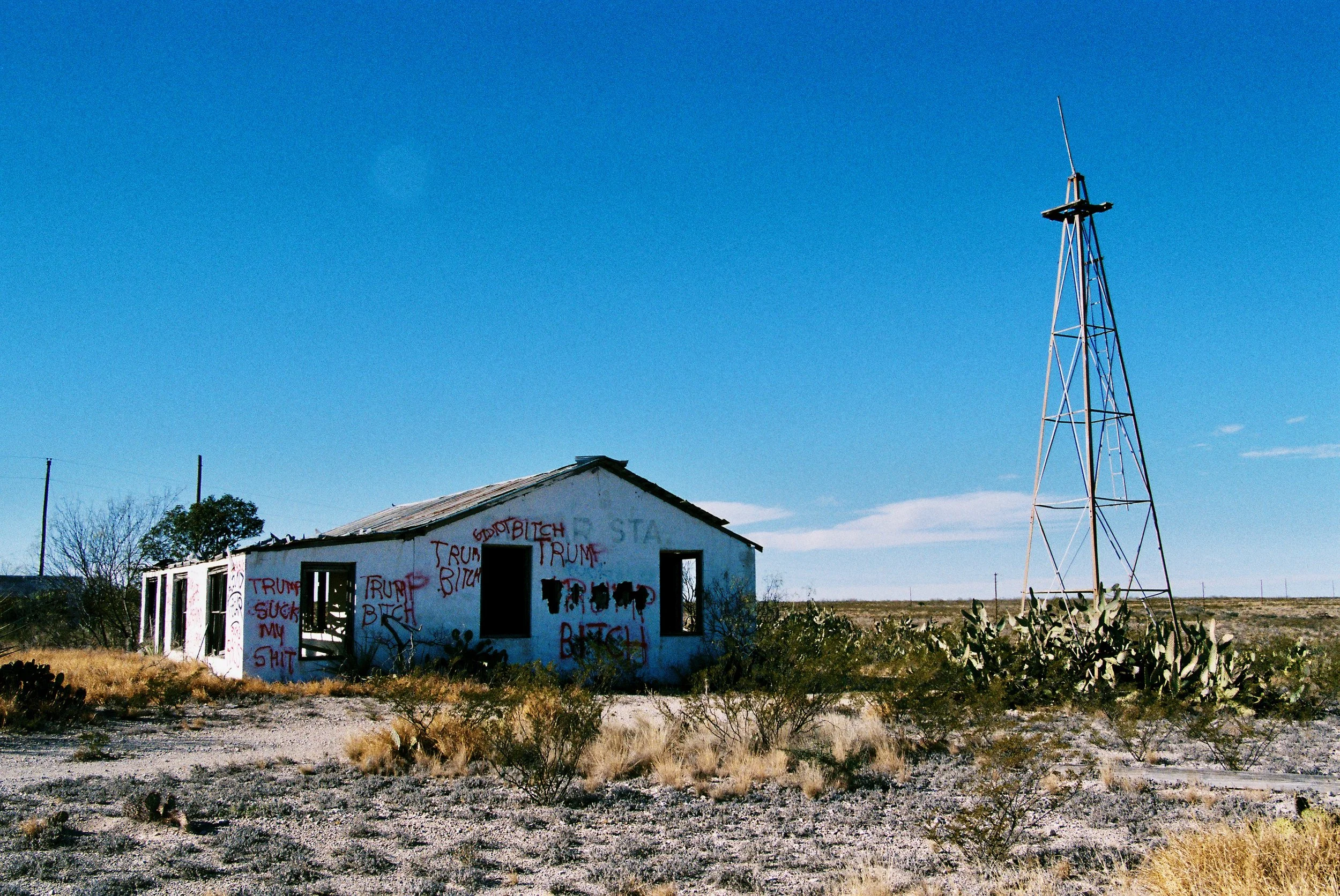 Abandoned building in a desert landscape with graffiti, including offensive language and political messages, and a tall metal tower to the right.