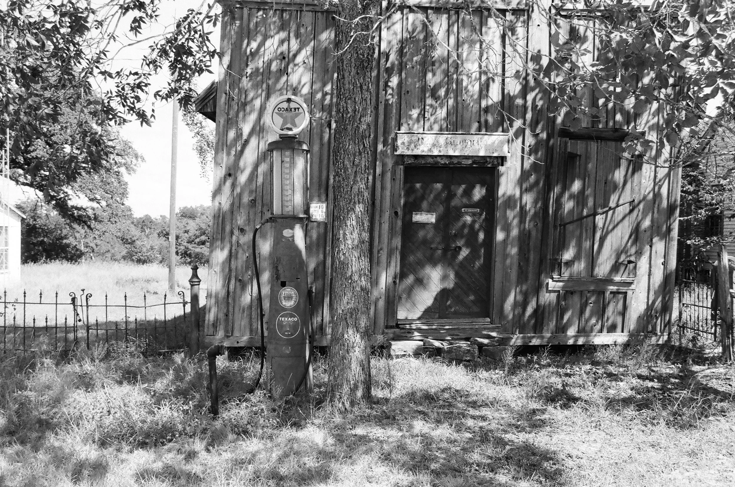 Black and white photo of a rustic wooden shed with a vintage gas pump outside, surrounded by trees and grass, with shadows cast by the leaves.