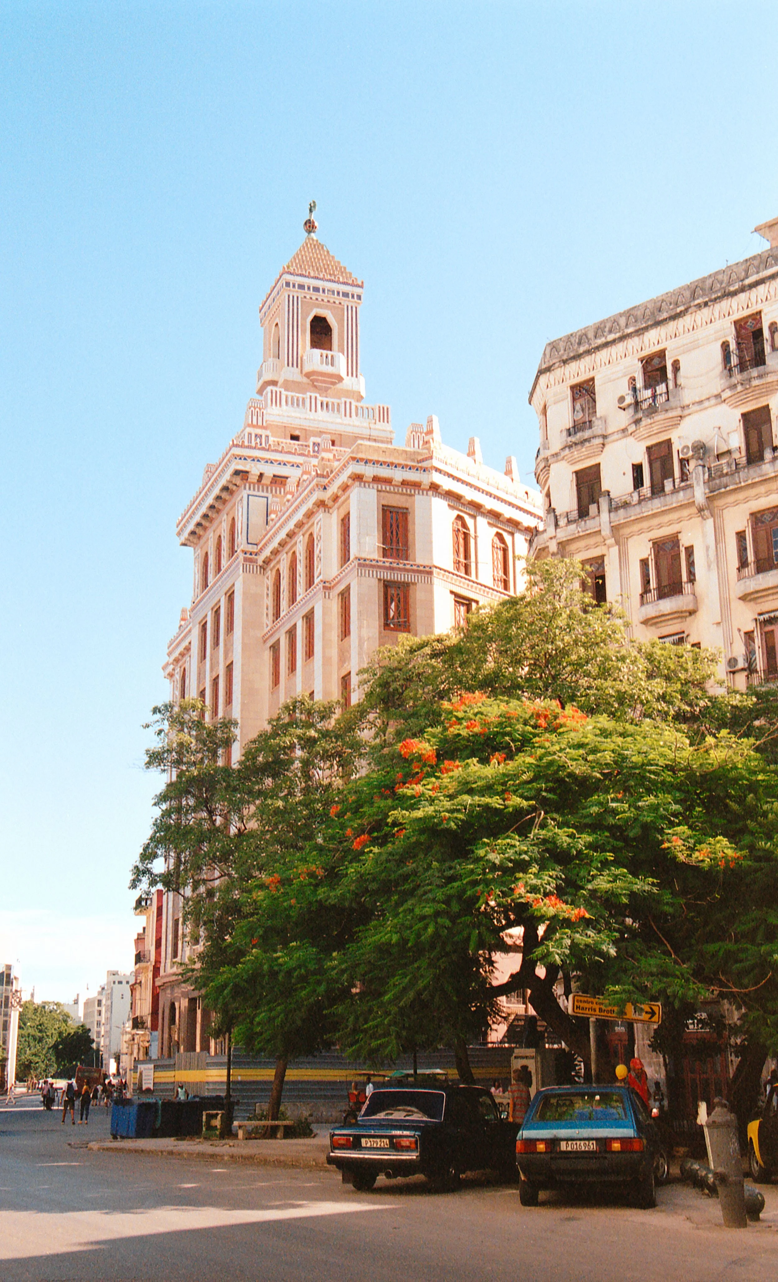 Street scene with historic buildings, trees, parked cars, and pedestrians under clear blue sky.