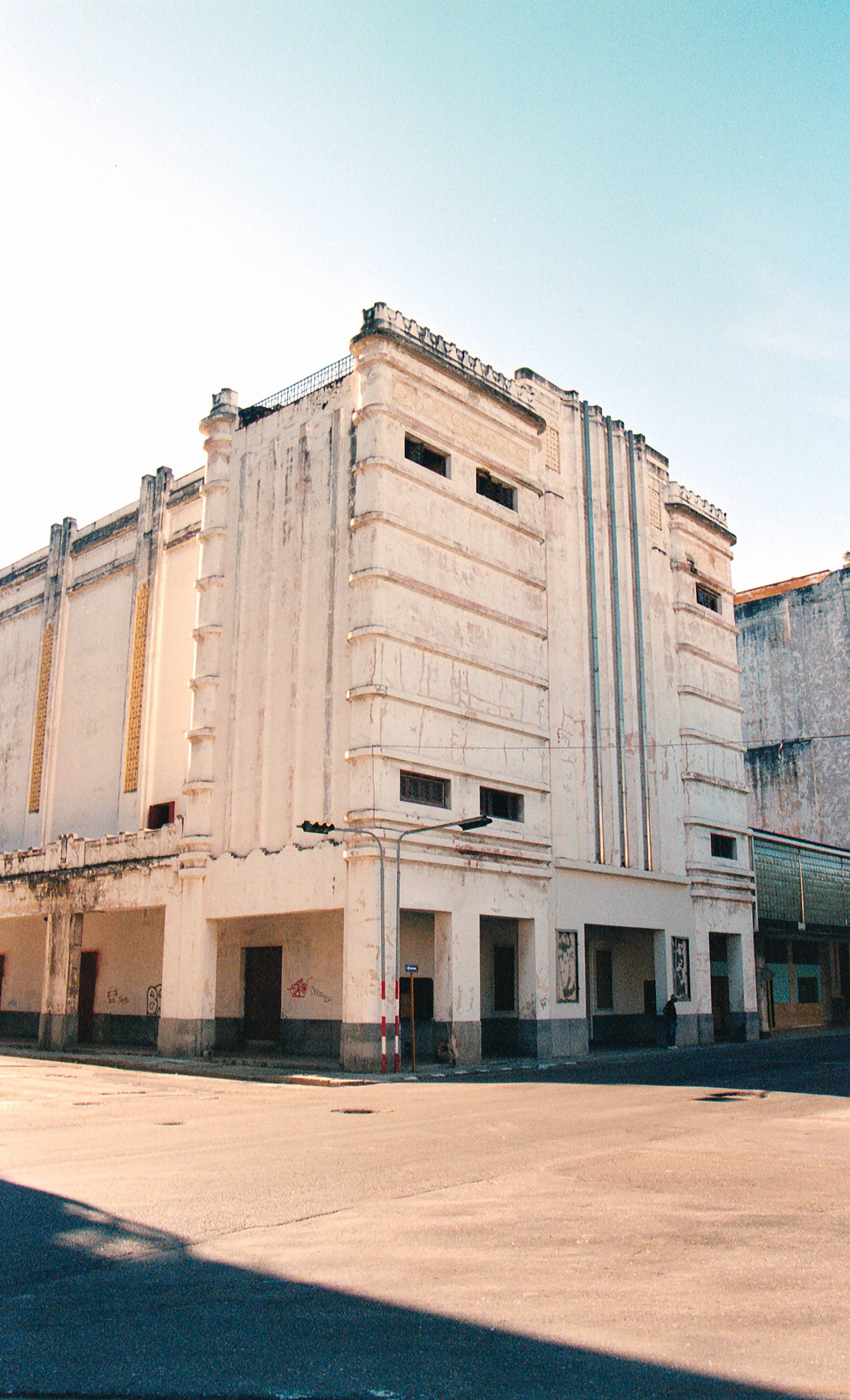 Old, weathered building with Art Deco architectural style, visible cracks, and minimal windows, located on a quiet street corner under a clear sky.