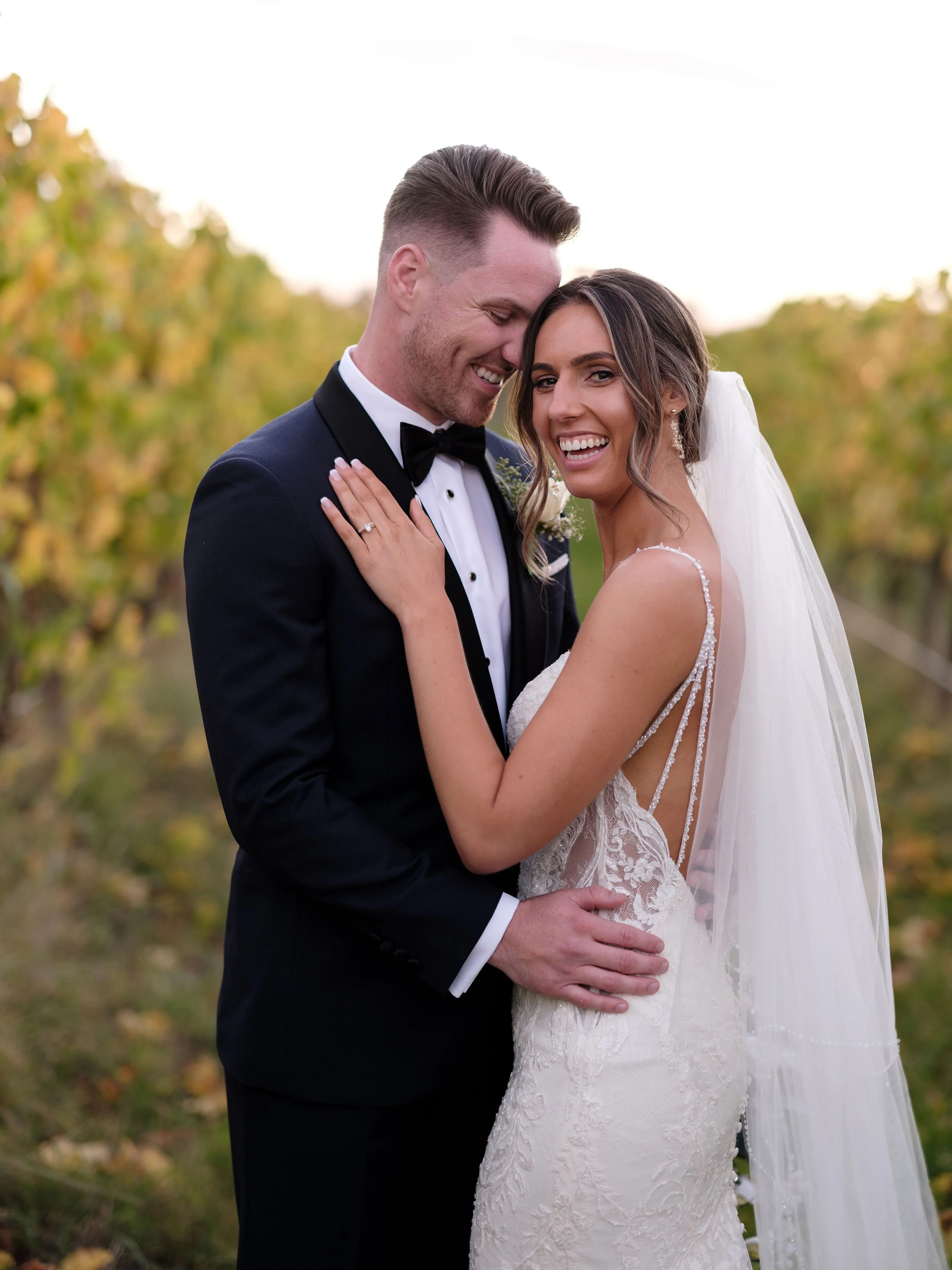 A newlywed couple, dressed in wedding attire, standing outdoors in a vineyard, smiling and embracing each other.