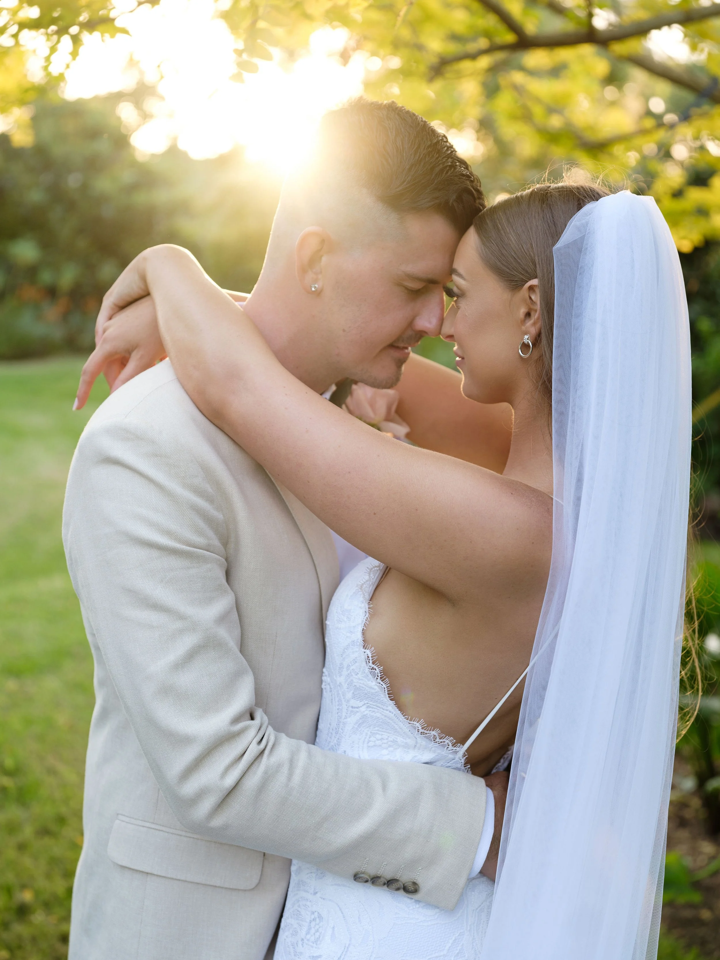 A bride and groom embrace outdoors during sunset, touching foreheads with eyes closed, in wedding attire.