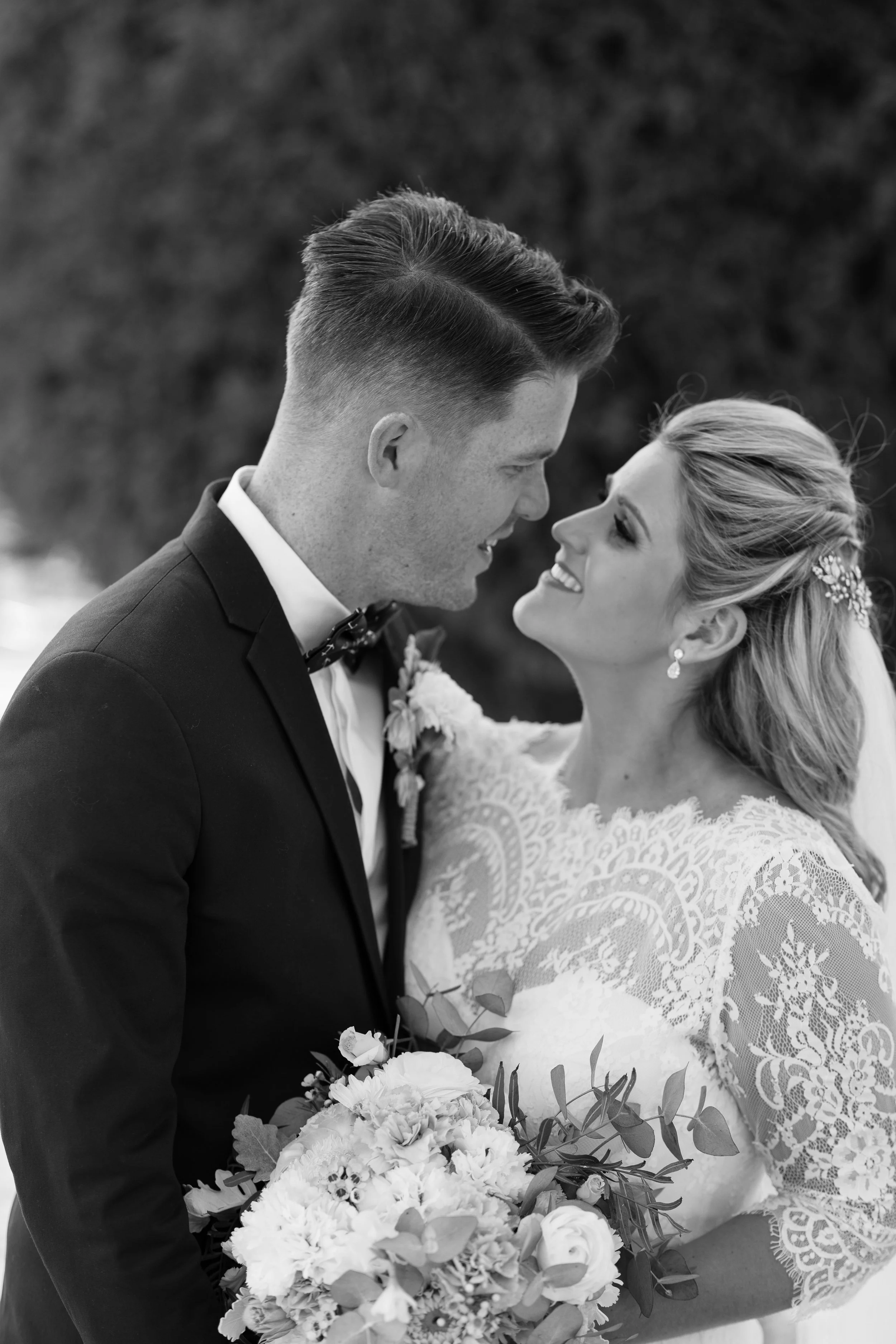 A black and white photo of a bride and groom smiling, facing each other closely. The bride is holding a bouquet of flowers and wearing a lace wedding dress, while the groom is dressed in a tuxedo with a bow tie. They appear joyful on their wedding day.