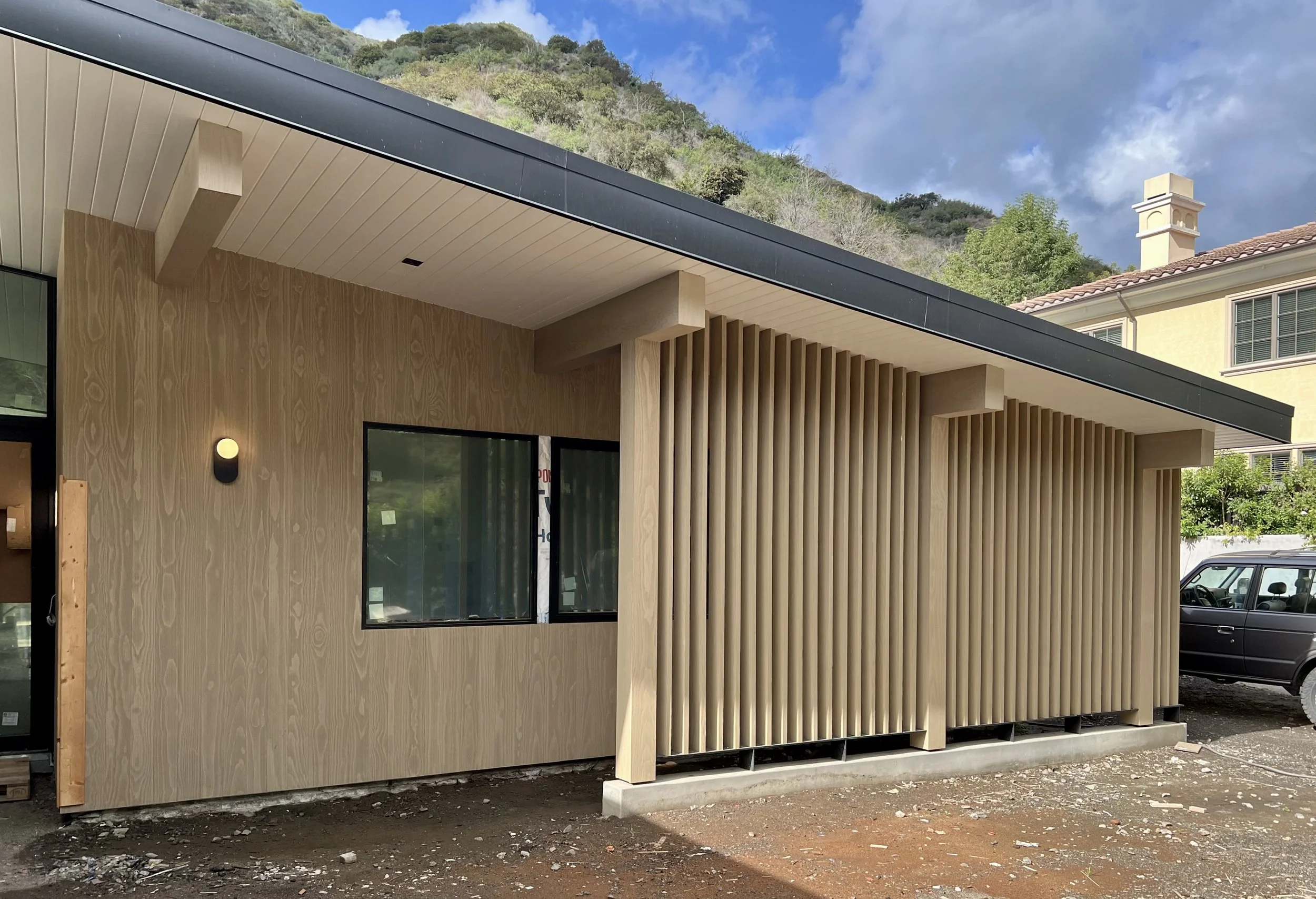 A modern building with wooden vertical slats on one side, a light wood exterior wall, a black window, and a black light fixture, with a hillside and a neighboring house in the background.