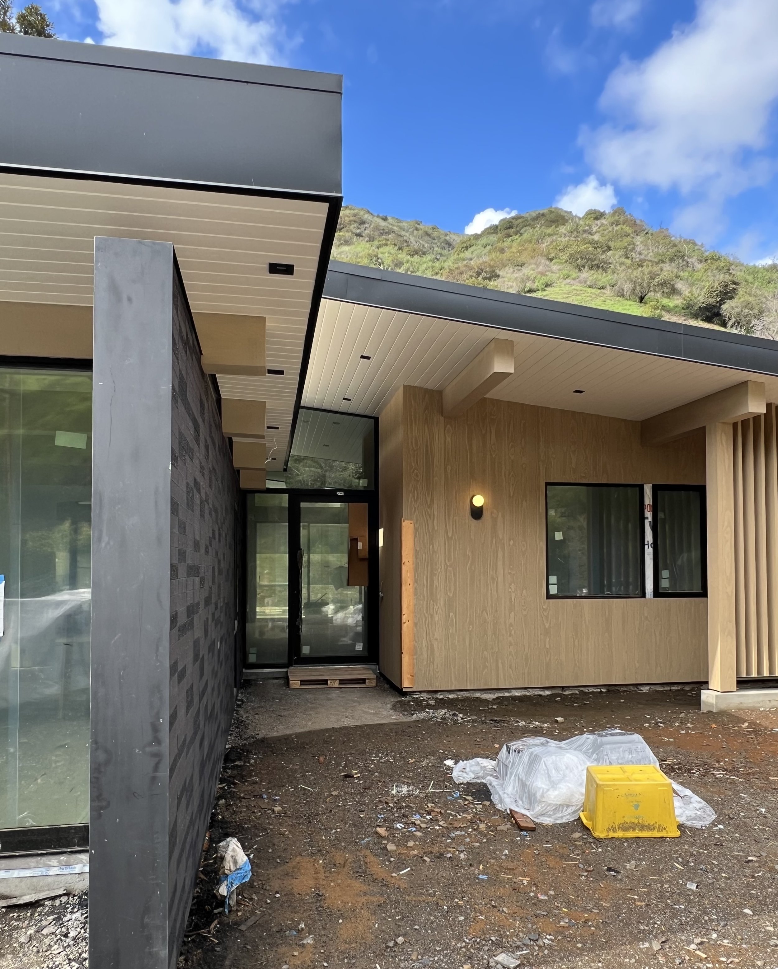 Construction site of a modern house with black, gray, and wood exterior panels, glass doors, and windows, with construction debris and equipment on the ground, and hills with trees in the background.