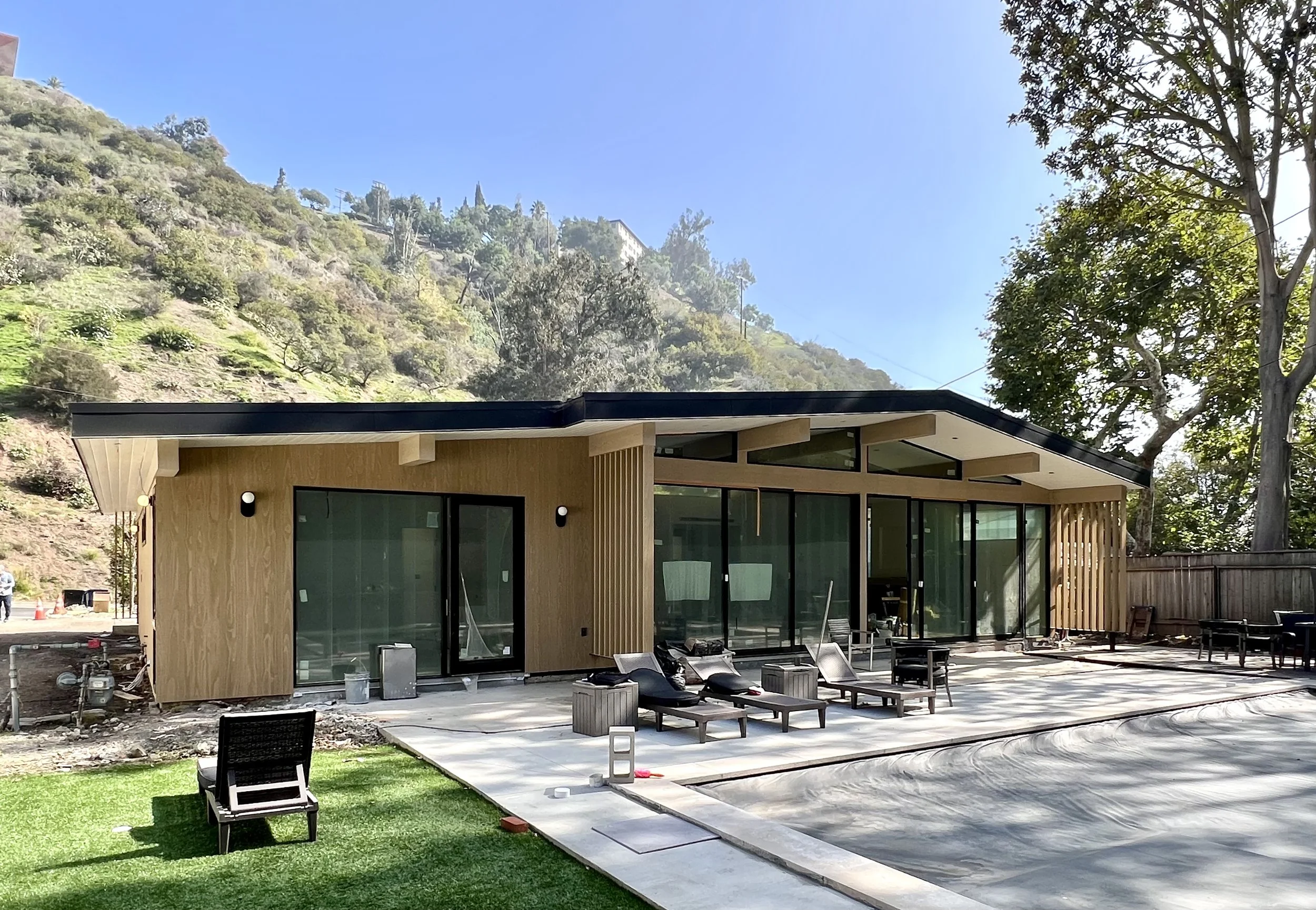 Modern house with large glass sliding doors and a sloped roof, surrounded by a backyard with lounge chairs, a grassy area, and a hillside with trees and plants in the background.