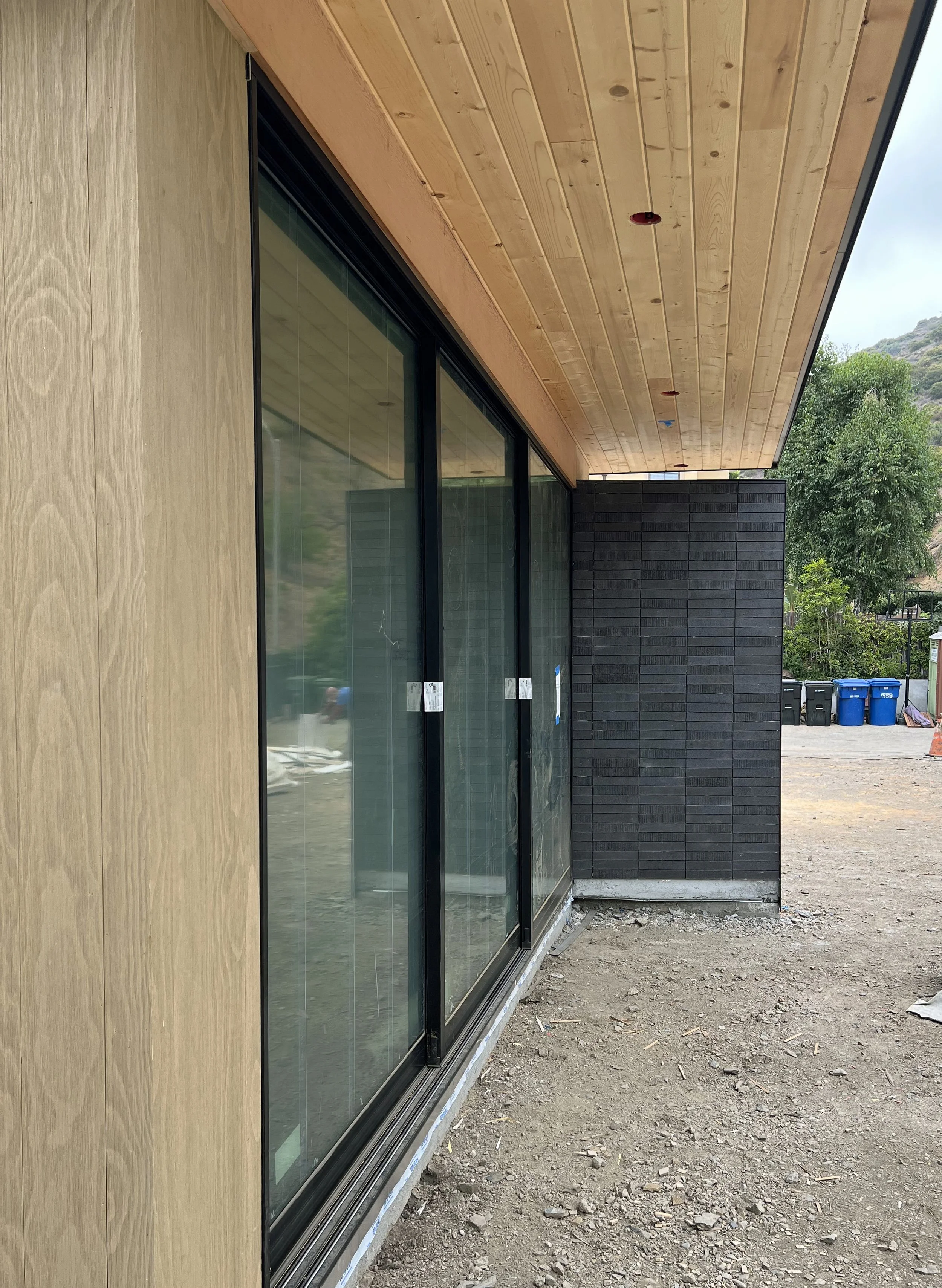 Exterior of a building under construction with large glass sliding doors, a wooden ceiling, and a black brick wall, with construction materials and trash bins in the background.