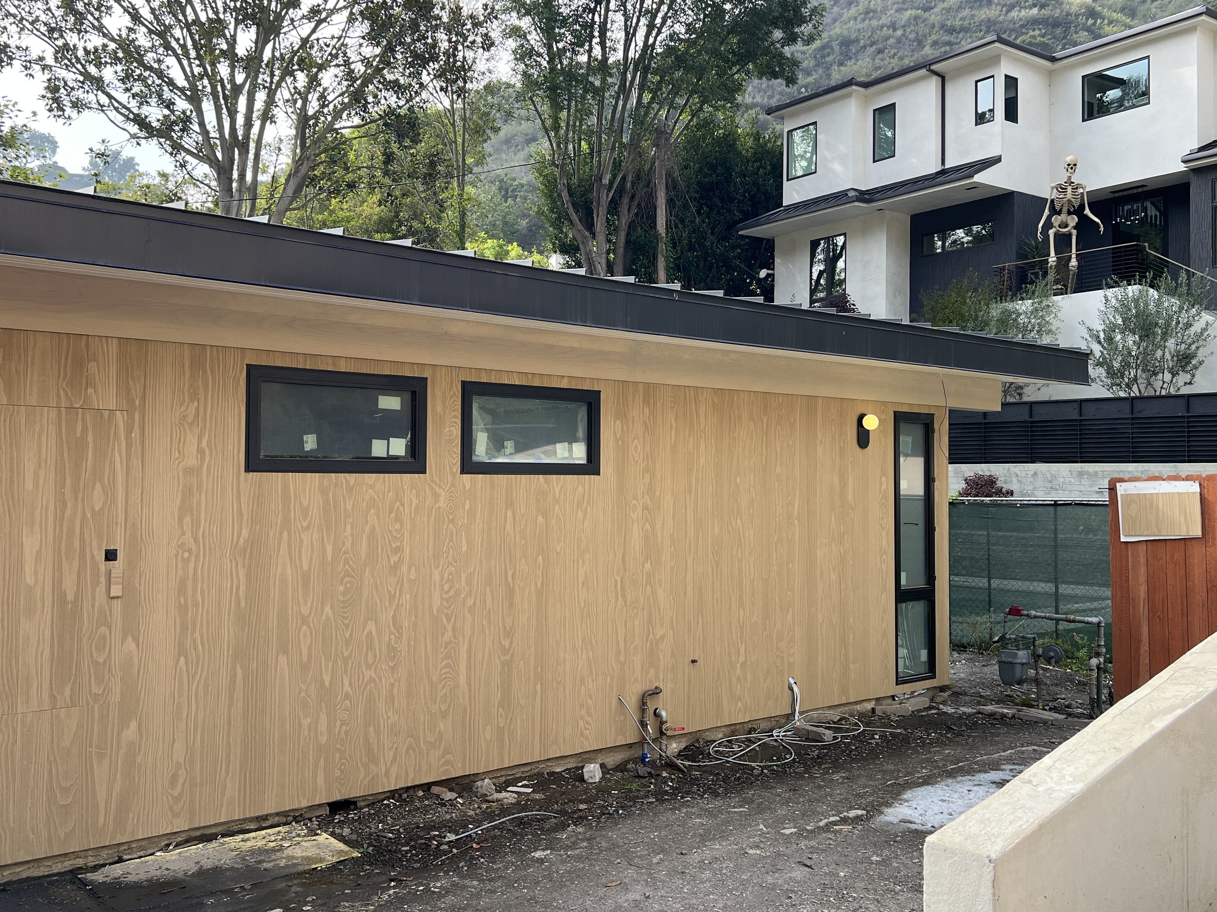 Construction site with a modern wooden building, two horizontal windows, and a door. A skeleton figure stands on the balcony of a white house in the background. Trees and hills are visible behind the buildings.