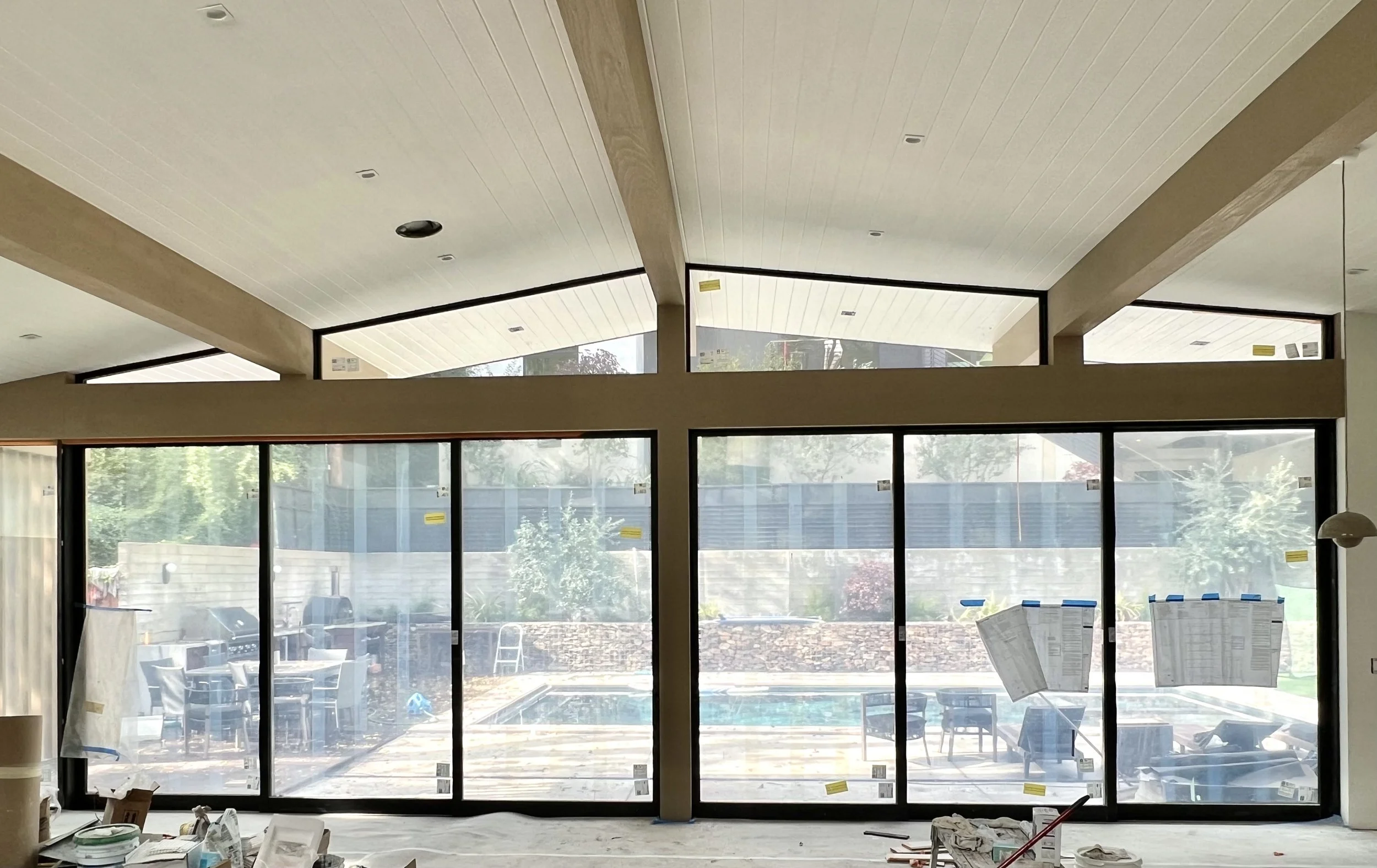 Interior view of a room under construction with large glass sliding doors leading to an outdoor patio with furniture and a pool, viewing into a backyard with trees and a fence.