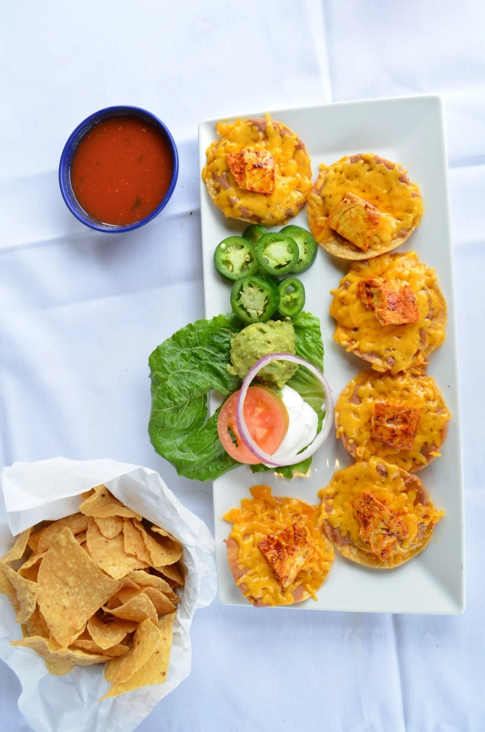 A plate of nachos topped with melted cheese and chunks of grilled chicken, sliced jalapeños, guacamole, shredded lettuce, sliced tomato and onion, served with a cup of salsa and a basket of tortilla chips on a white tablecloth.