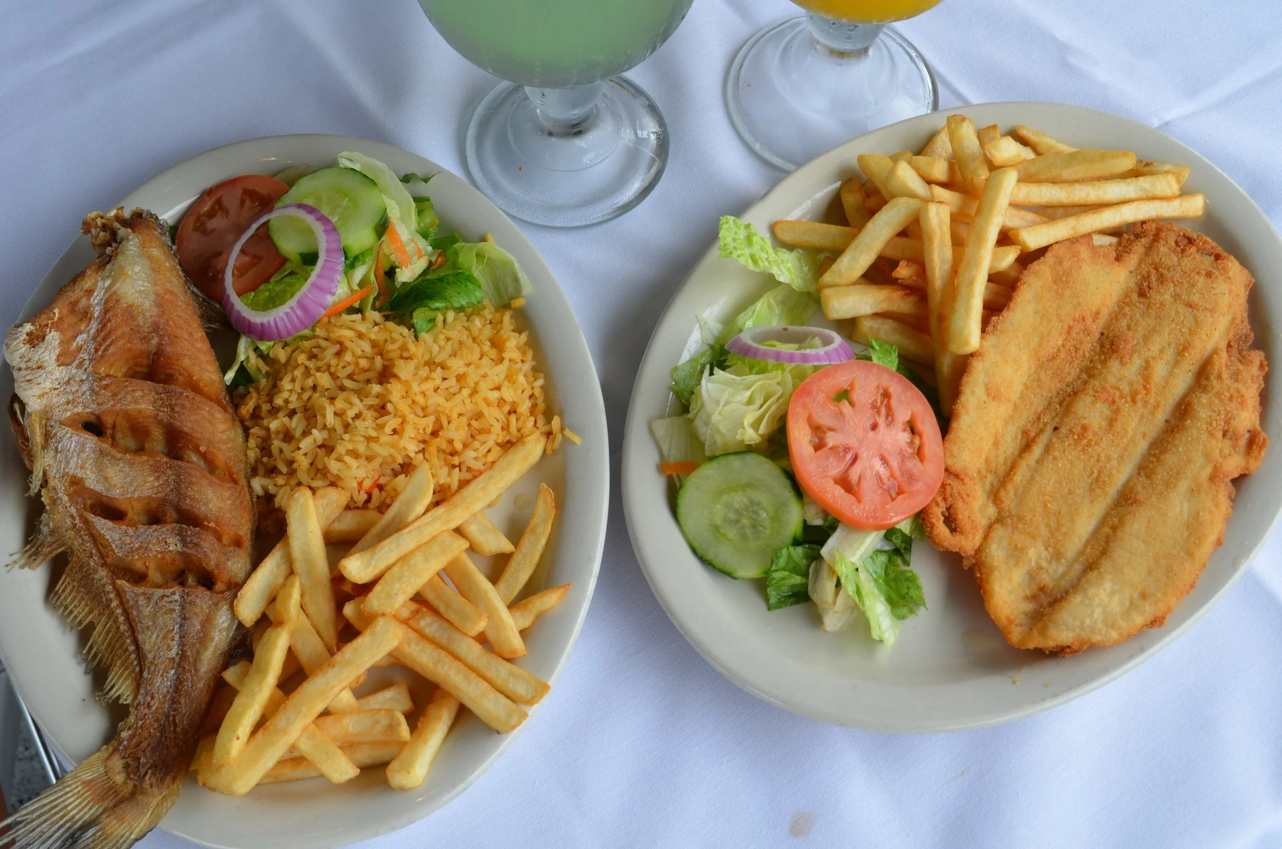 Two plates of fried fish, French fries, and salad with lettuce, tomato, cucumber, and onion, with two glasses of drinks in the background on a white tablecloth.