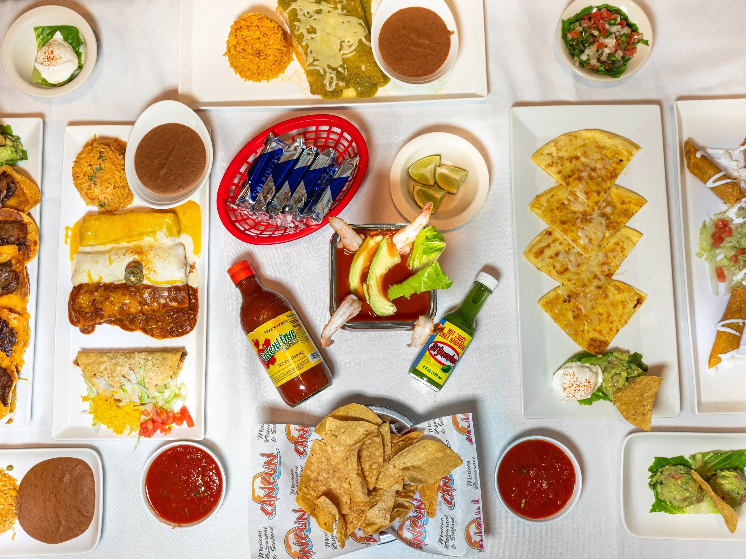 A variety of Mexican dishes on white plates, including nachos, tacos, Mexican rice, and salsa, along with bottles of hot sauce and beverages, on a white table.