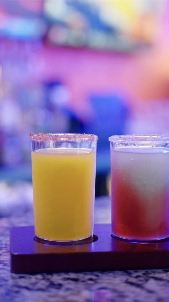Two clear glasses filled with colorful beverages, one yellow and one pink, placed on a wooden tray on a bar or counter with a blurred, colorful background.