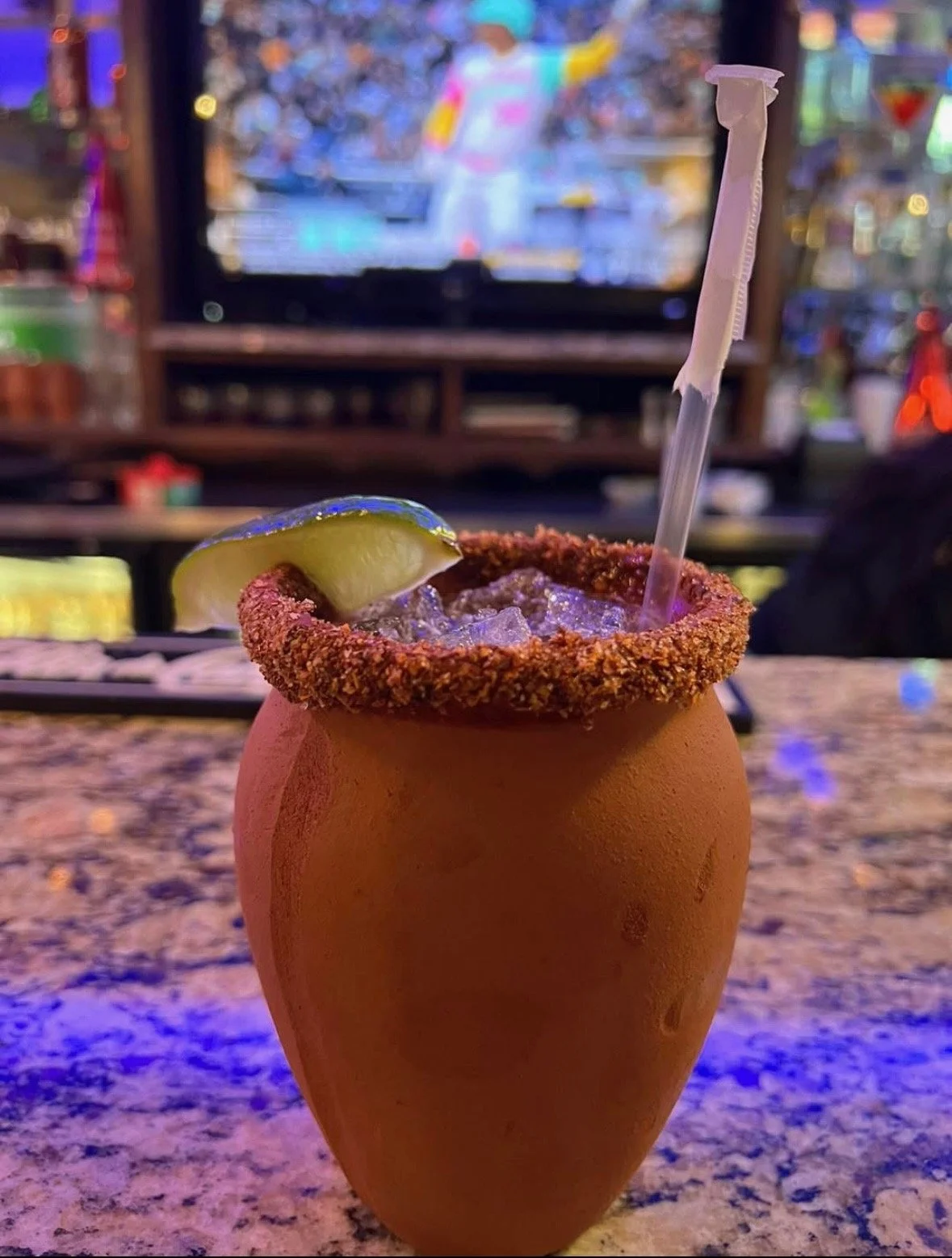 A cocktail in a terracotta-style clay cup with a chili rim, lime wedge, ice, and a straw, sitting on a bar counter with a blurred TV screen and bar background in available lighting.