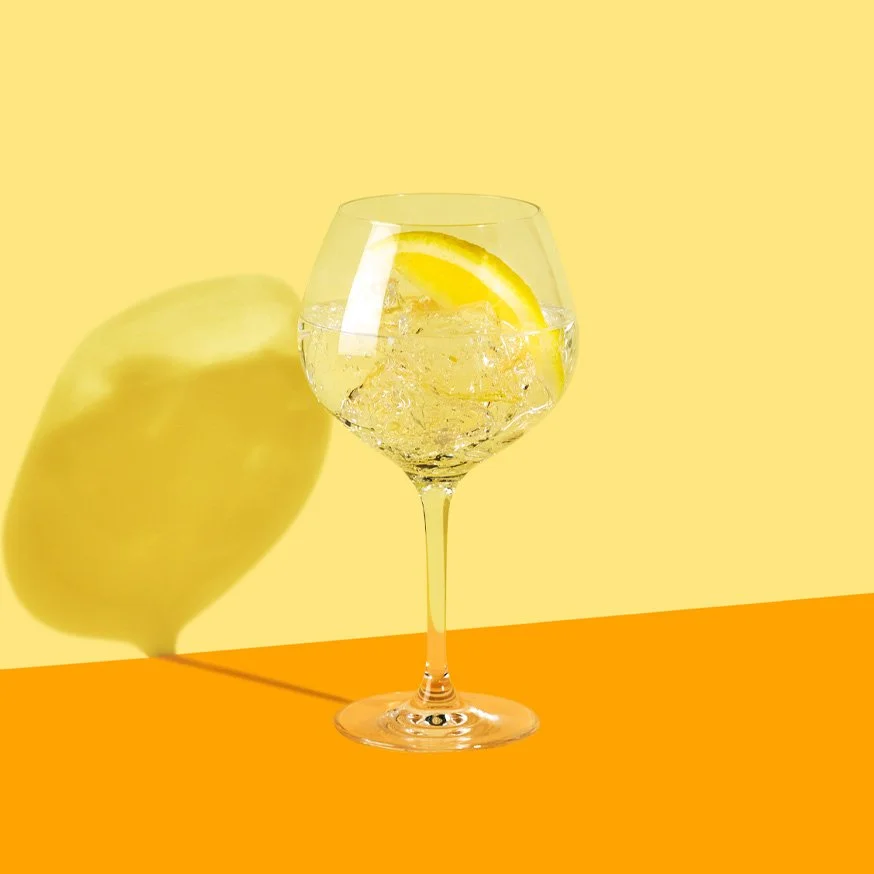 Gin och tonic med fläder och citron i ballongglas mot gul bakgrund.